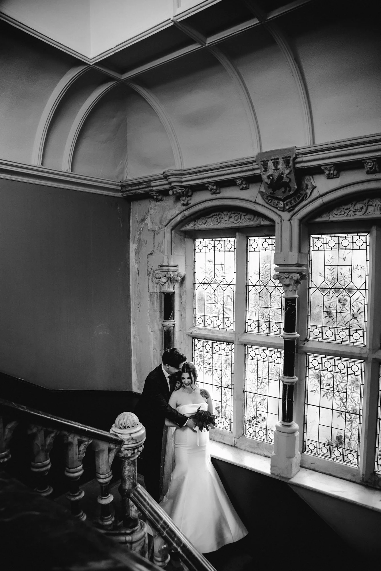 A black and white photograph of a bride and groom standing on a staircase inside a historic building, near large stained glass windows. The bride is wearing a strapless wedding gown holding a bouquet, and the groom is in a dark suit, gently leaning t