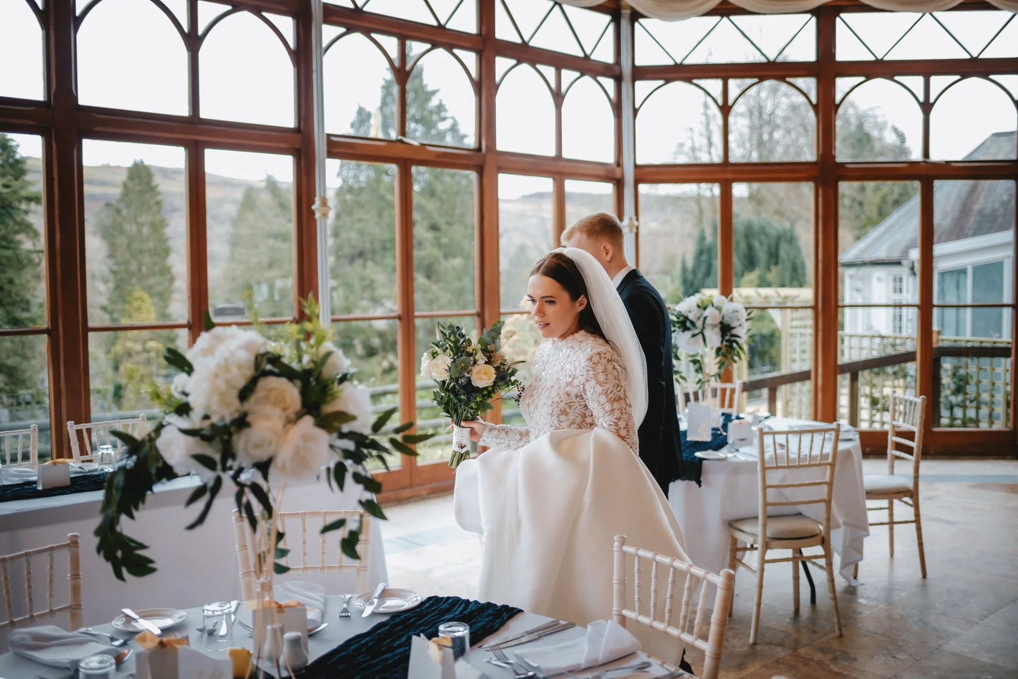 A bride in a white wedding dress holding a bouquet standing next to a groom in a black suit inside a glass-enclosed venue with wooden framing and decorated tables.