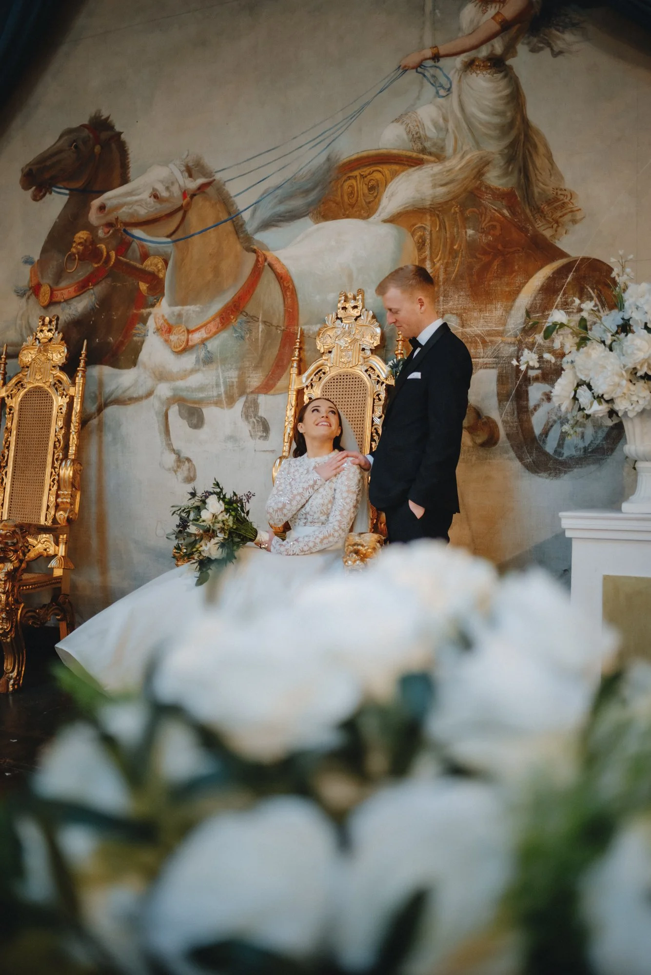 A bride sitting on a golden throne in a wedding dress looking up at a groom standing beside her, in a luxurious setting with a large, detailed mural of a horse-drawn carriage in the background.