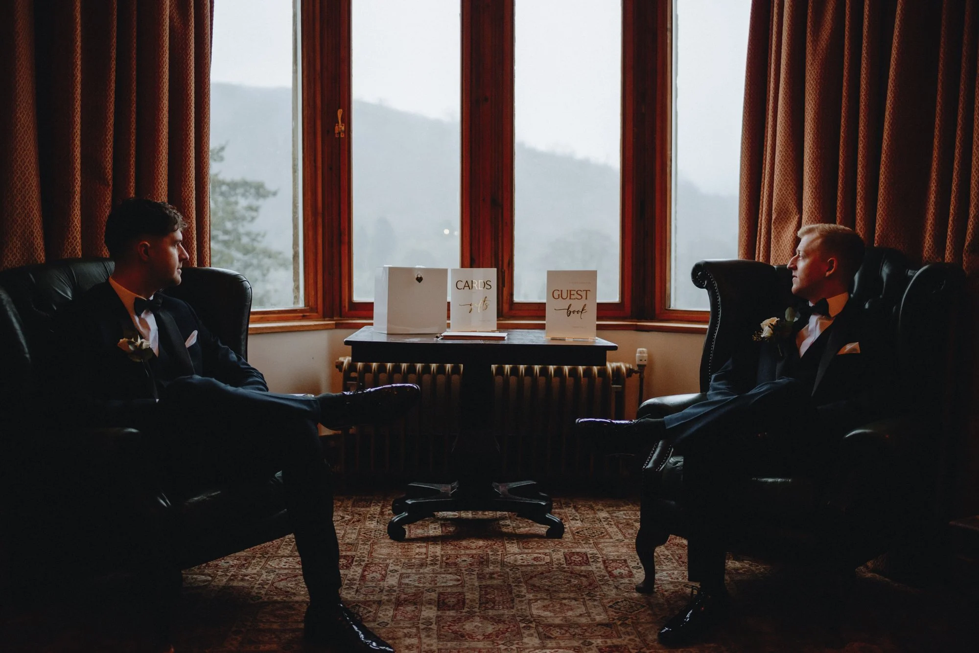 Two men in tuxedos facing each other while sitting on black leather armchairs in front of a large window with views of a foggy landscape, with wedding signs on a table between them.