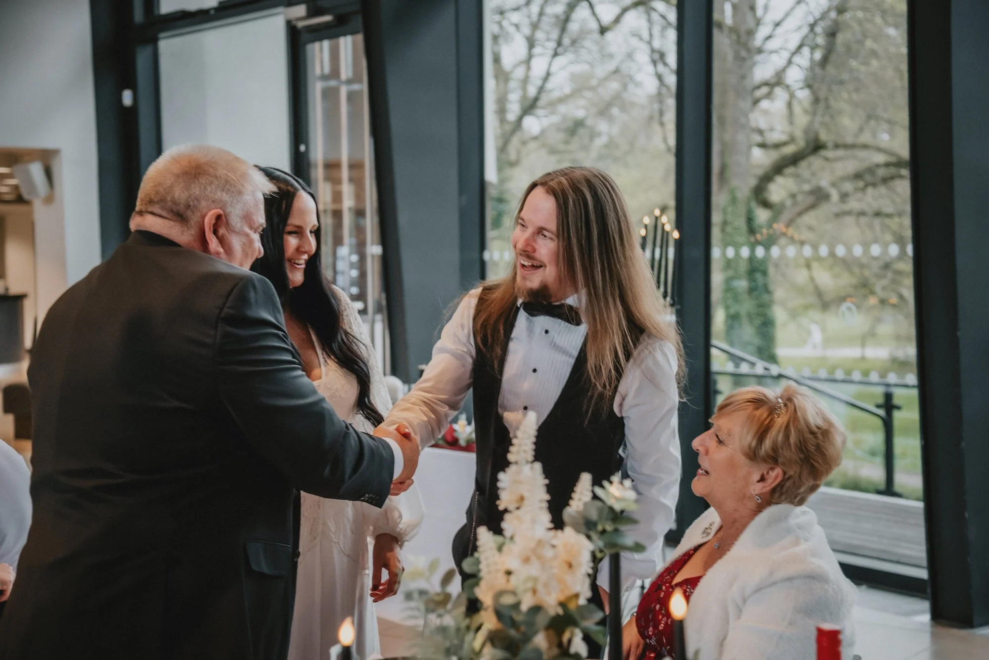 People dressed formally at a celebration, with two men and two women smiling and shaking hands inside a decorated venue with large windows and a view of trees outside.