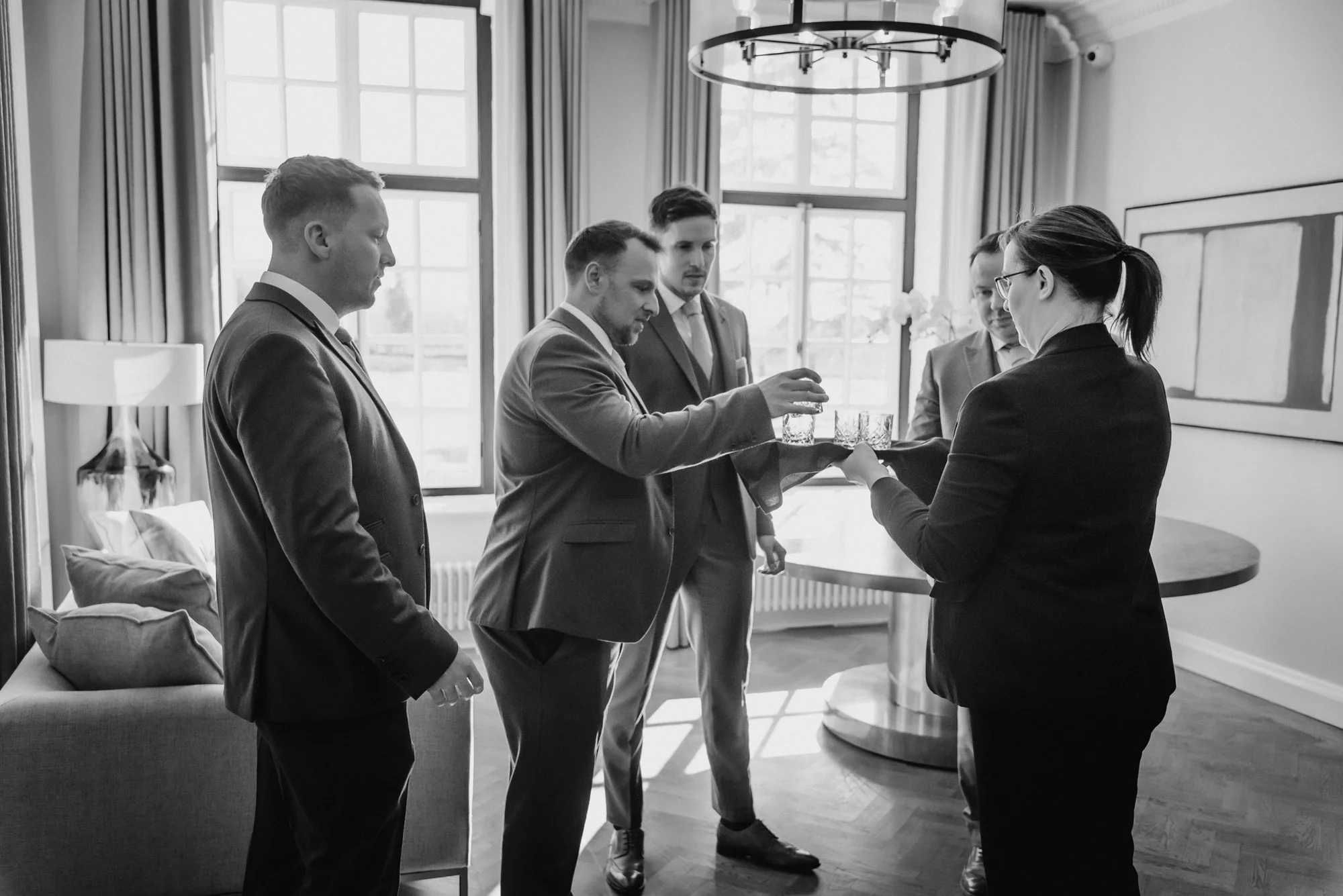 Five people in professional attire participating in a formal indoor event, with three men and two women, as one woman serves drinks from a tray to one of the men.