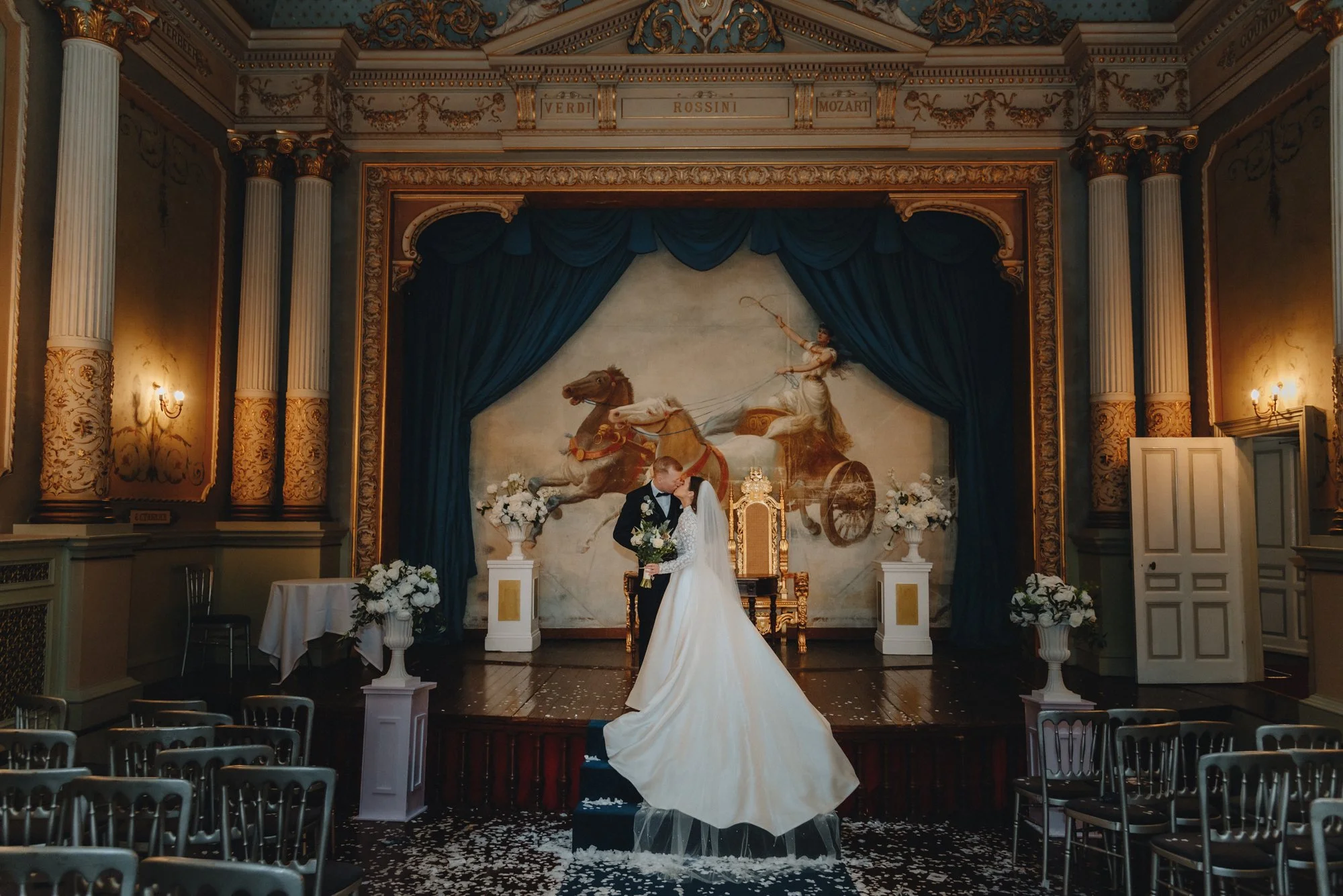 A bride and groom kissing on a small stage during their wedding ceremony, with a painted backdrop of a chariot pulled by horses, surrounded by floral arrangements and draped curtains in an ornate hall.