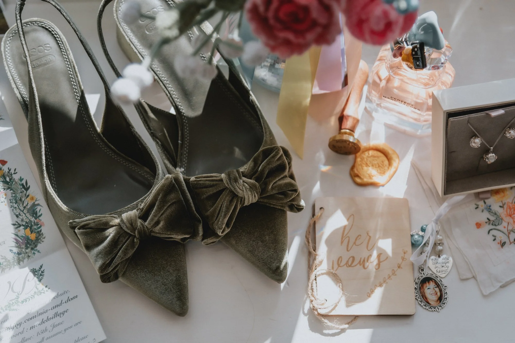 A display of jewelry, a pair of memory shoes with bows, a floral candle, perfume bottles, a greeting card that says 'her vents,' and a jewelry box with a necklace and earrings, arranged on a white surface.