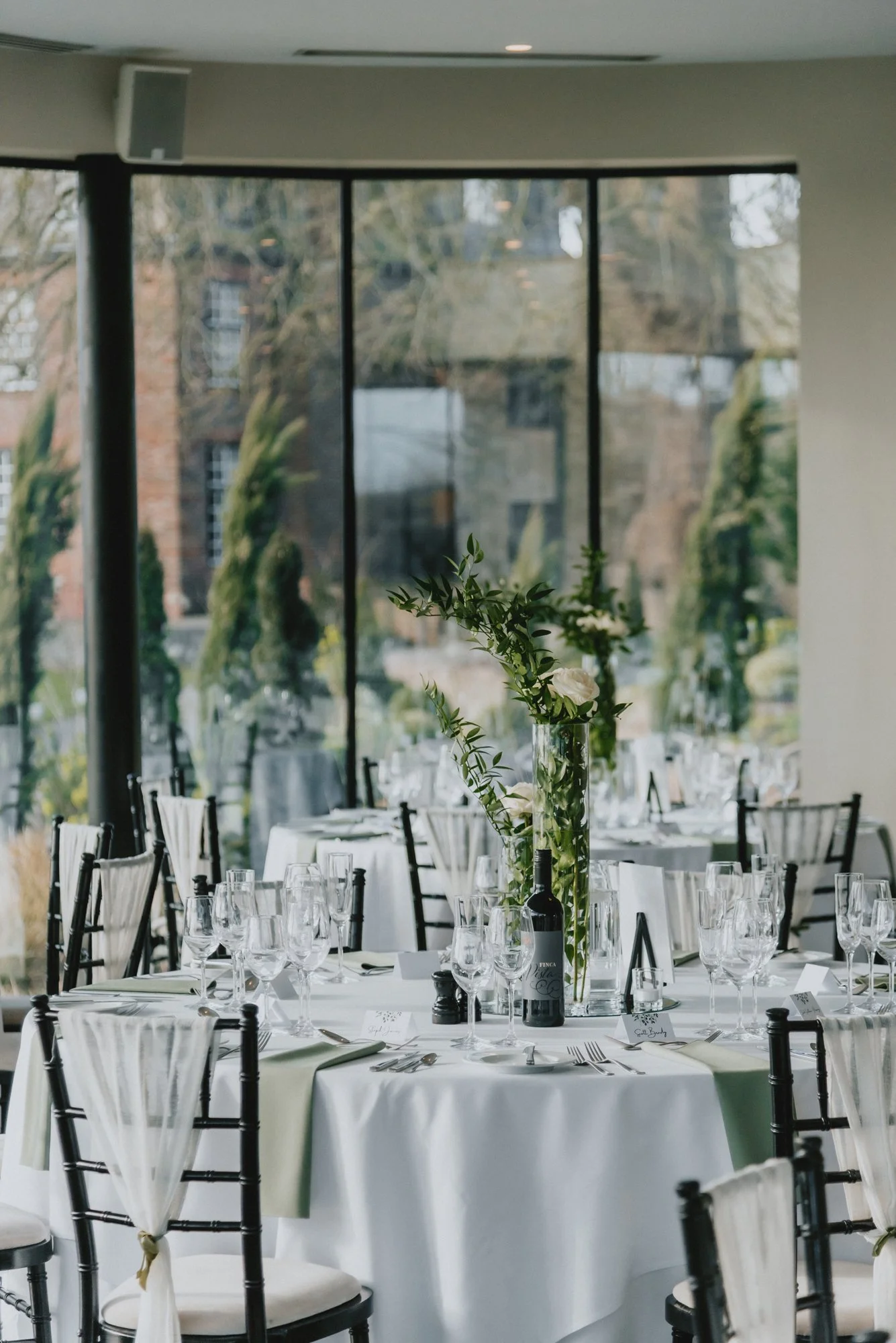 Elegant banquet table set with white tablecloth, glassware, silverware, and a tall floral center with greenery and white flowers, in a modern venue with large windows.