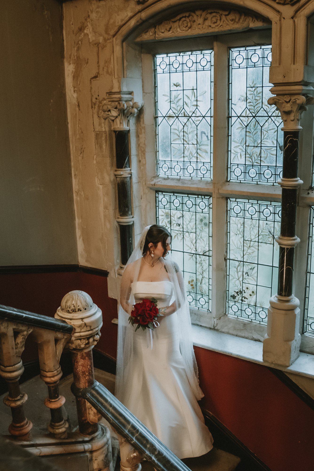 A bride in a white wedding dress holding a bouquet of red roses, standing on a staircase near an ornate stained glass window with floral designs, in a historic building.