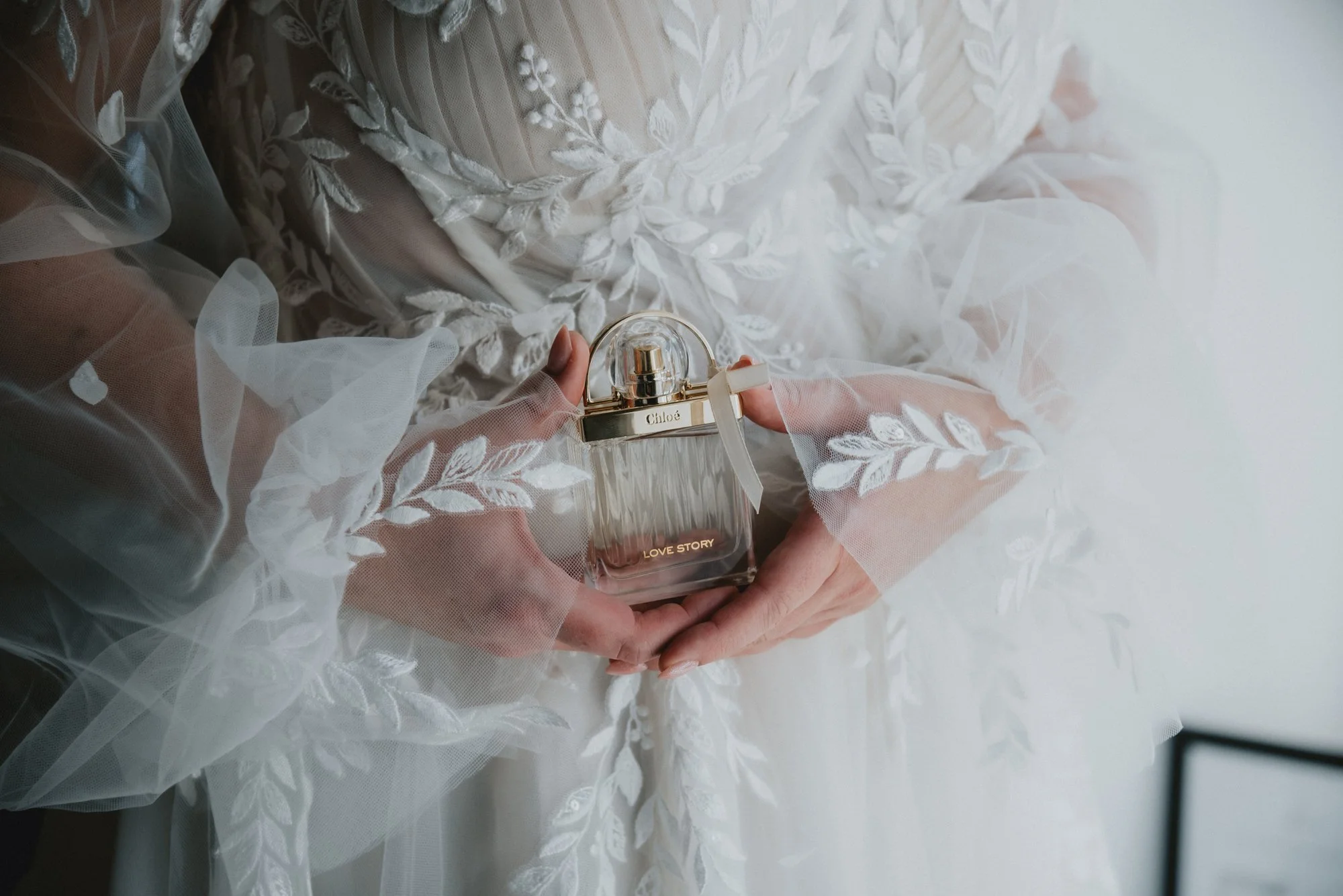A woman in a white dress with floral embroidery holds a perfume bottle labeled 'Love Story' by Chloé, with lace and delicate fabric details visible.