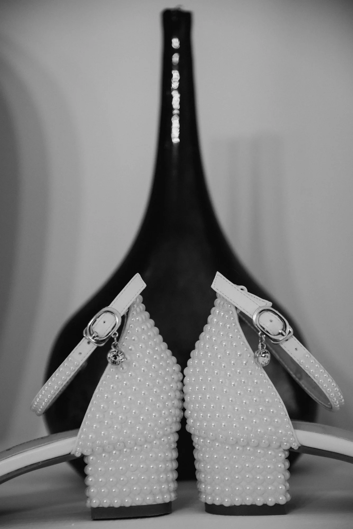 A pair of white high-heeled shoes decorated with pearls, placed on a surface with a dark vase in the background.