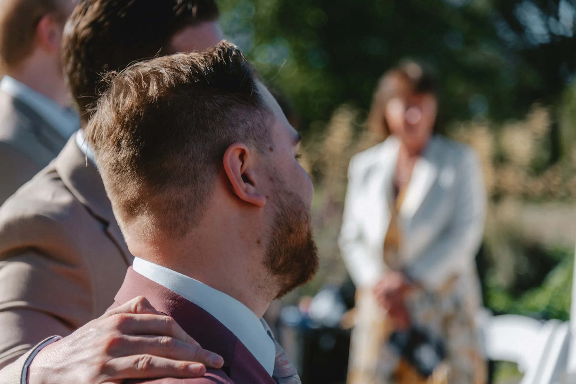 People gathered outdoors, with the focus on a young man with short brown hair and a beard, sitting with others. A woman in a light-colored blazer is blurred in the background.