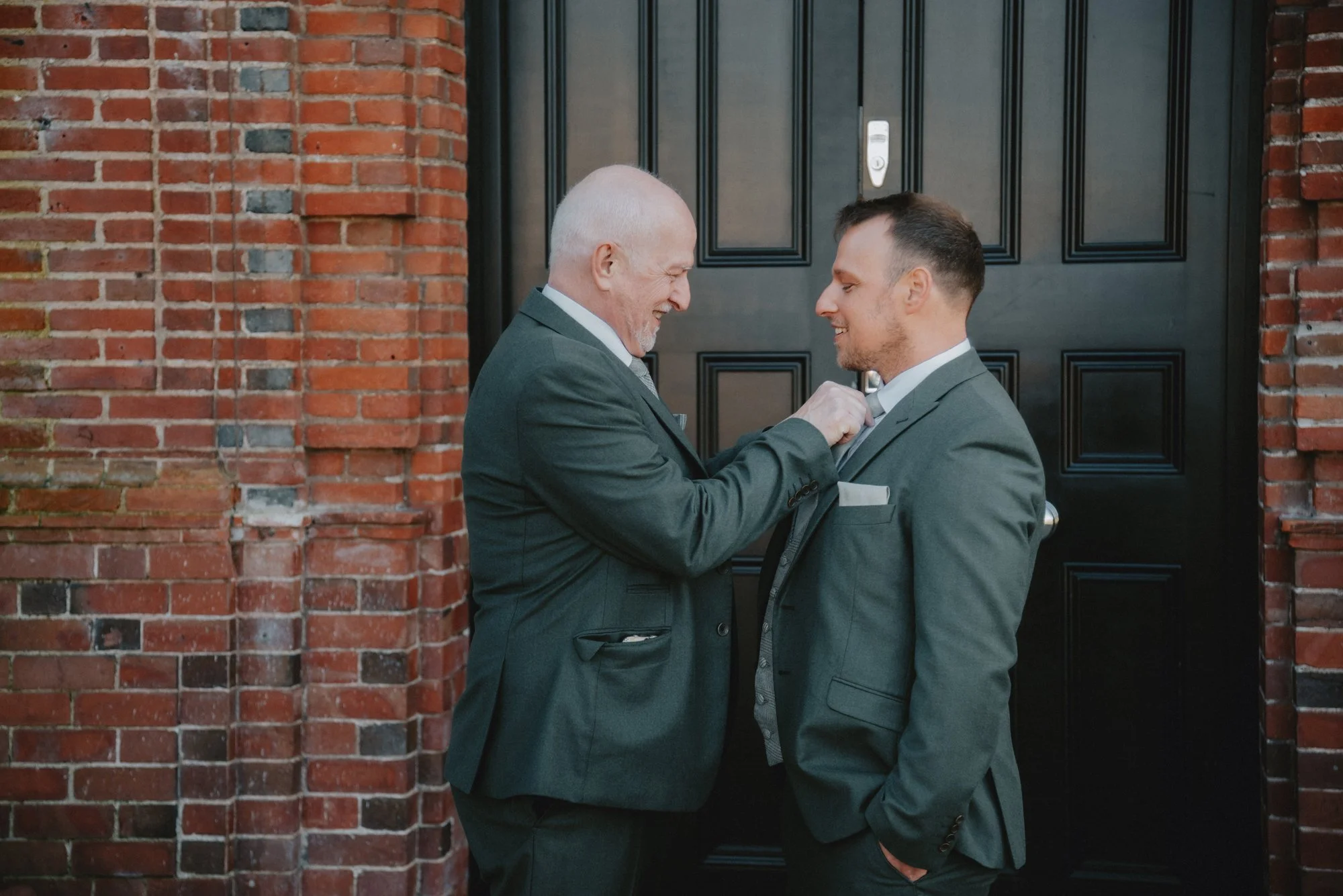 An older man and a younger man in suits smiling at each other in front of a black door and brick wall, celebrating a special occasion.