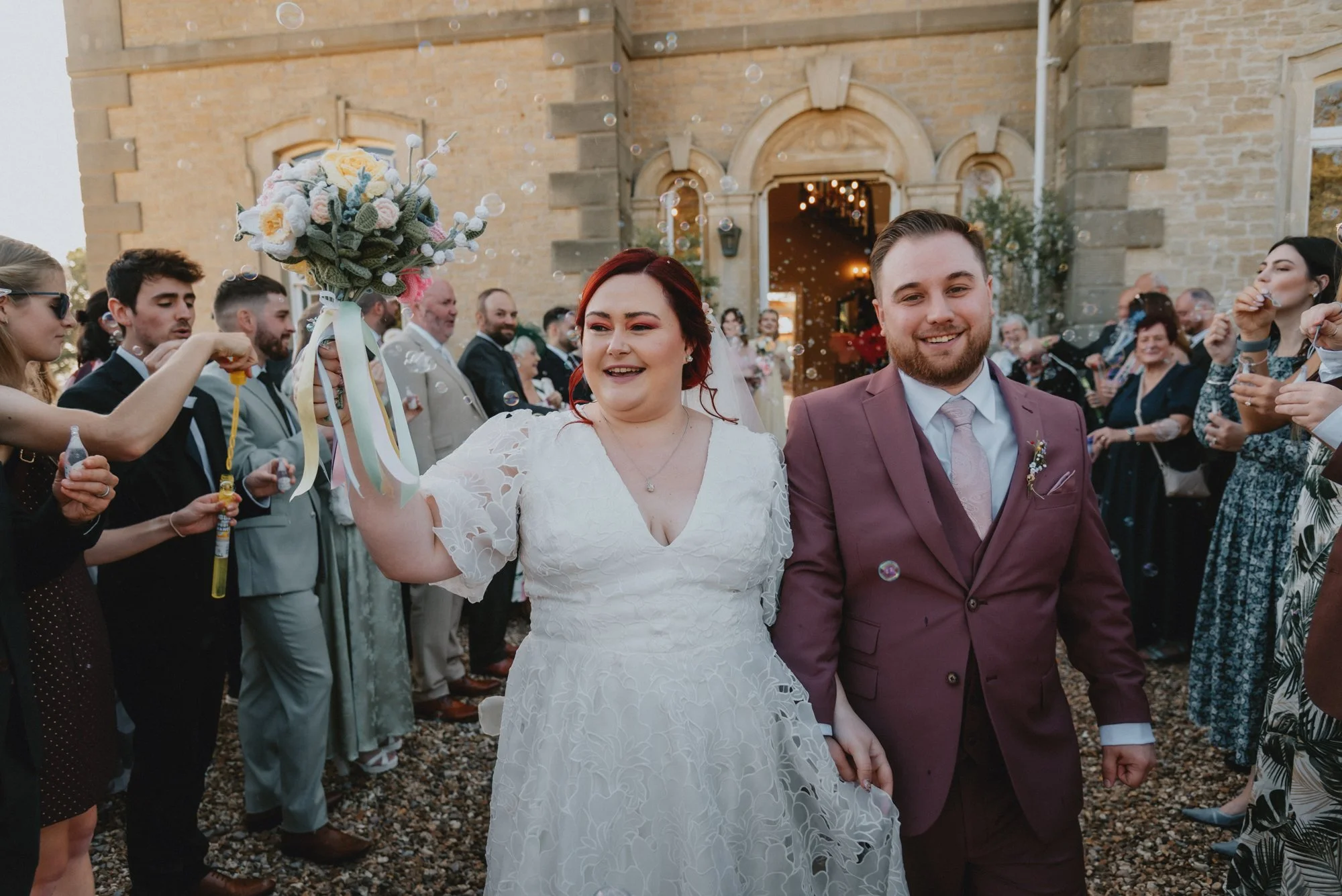 A newlywed couple holding hands and smiling, walking through a crowd outside a church during their wedding celebration. The bride is holding a bouquet and wearing a white lace dress, while the groom is in a maroon suit. Guests are celebrating and blo