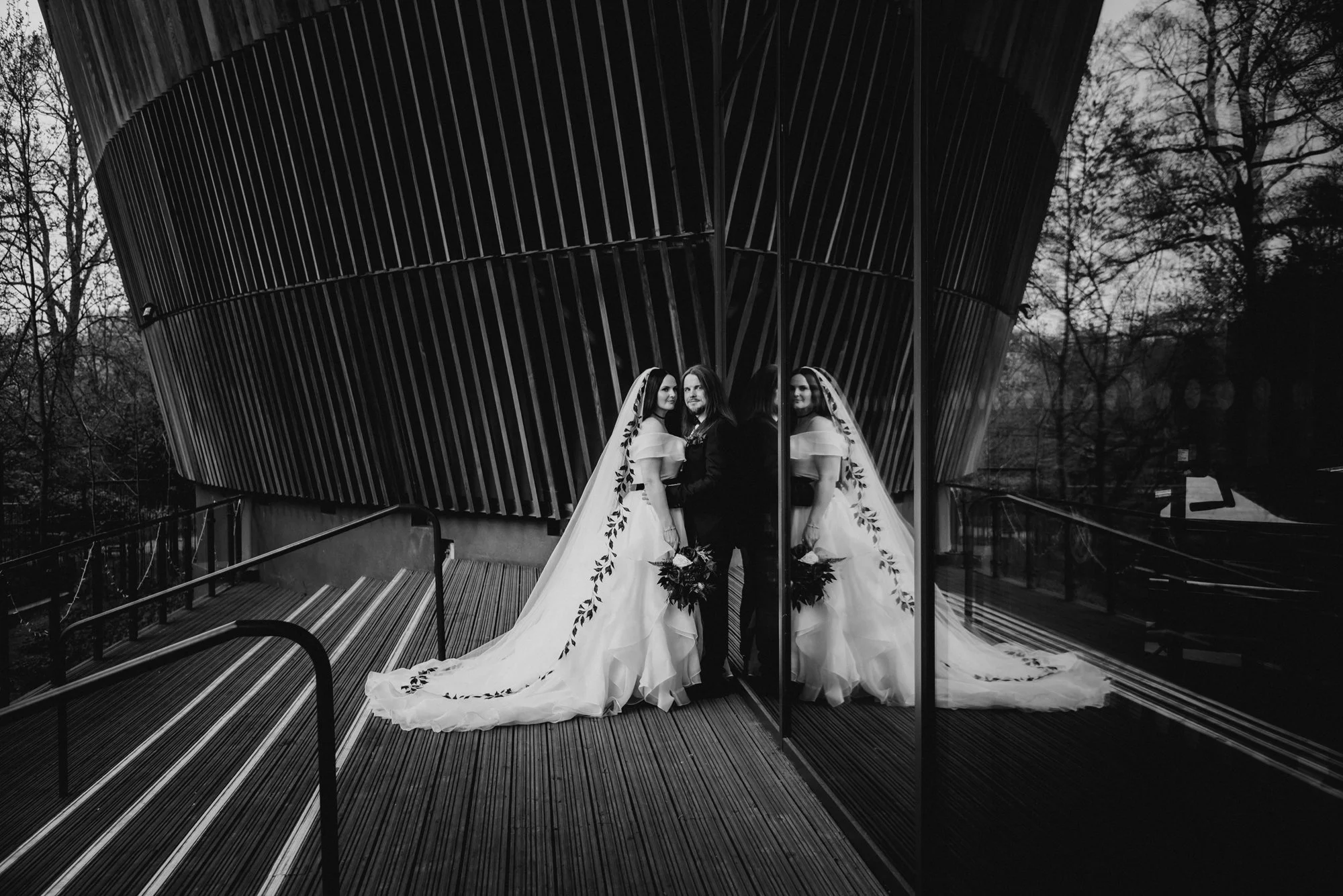 Black and white photo of a bride and groom standing side by side, holding hands, with a mirrored wall reflecting their image. The bride is wearing a wedding dress with a long train and veil, while the groom is in a dark suit, on a modern outdoor stai