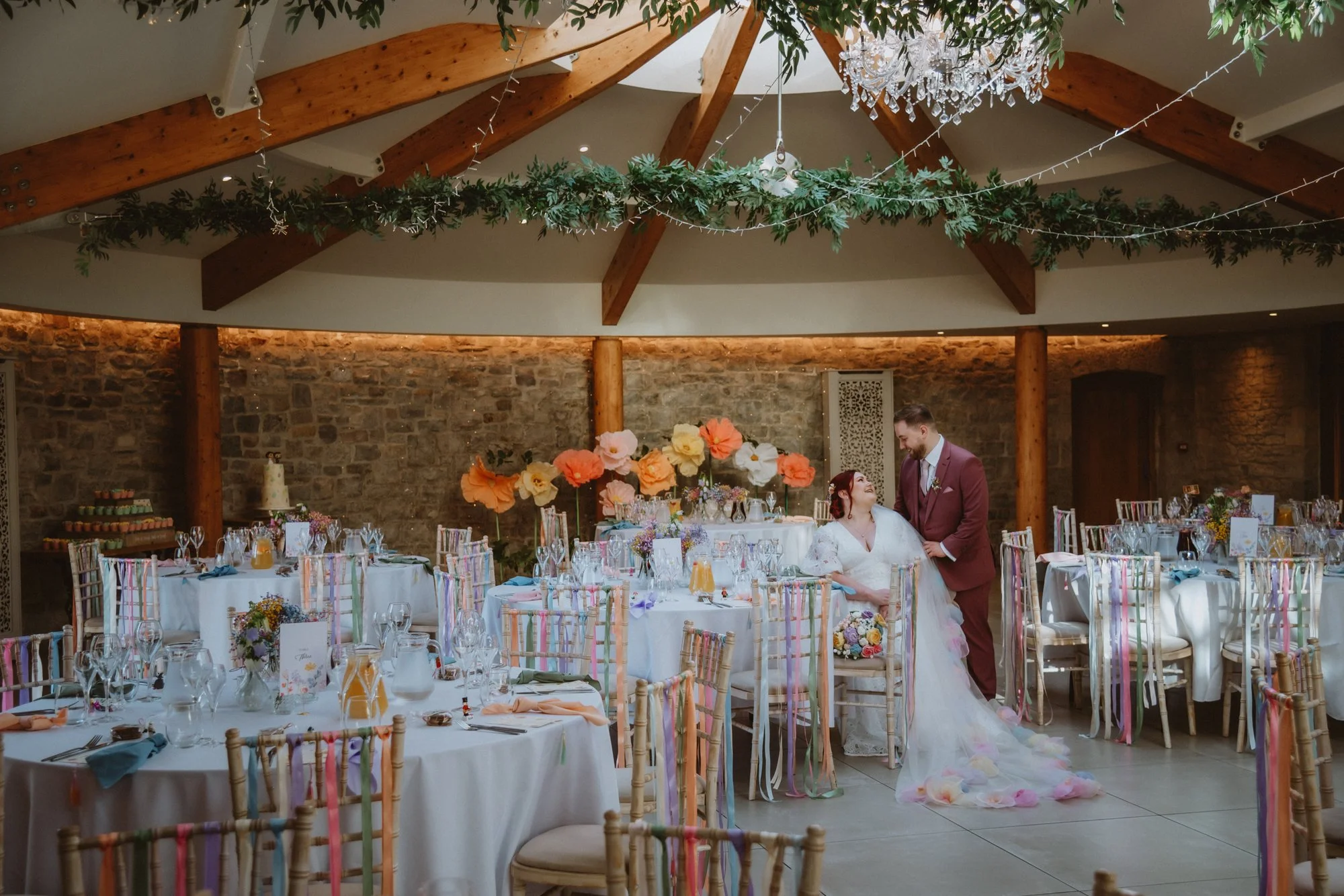 A wedding reception with a bride and groom standing together in a decorated banquet hall. The bride is dressed in a white wedding gown and the groom in a maroon suit. The tables are set with glassware, napkins, and centerpieces. Large colorful paper 