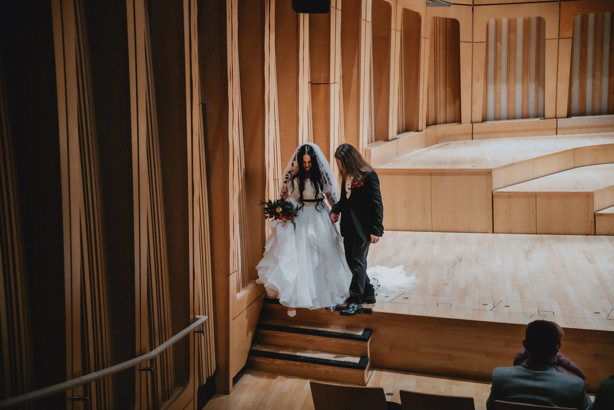 A bride in a white wedding gown holding a bouquet and a woman in a black suit walking on stage in a wooden auditorium, with a few seated audience members watching