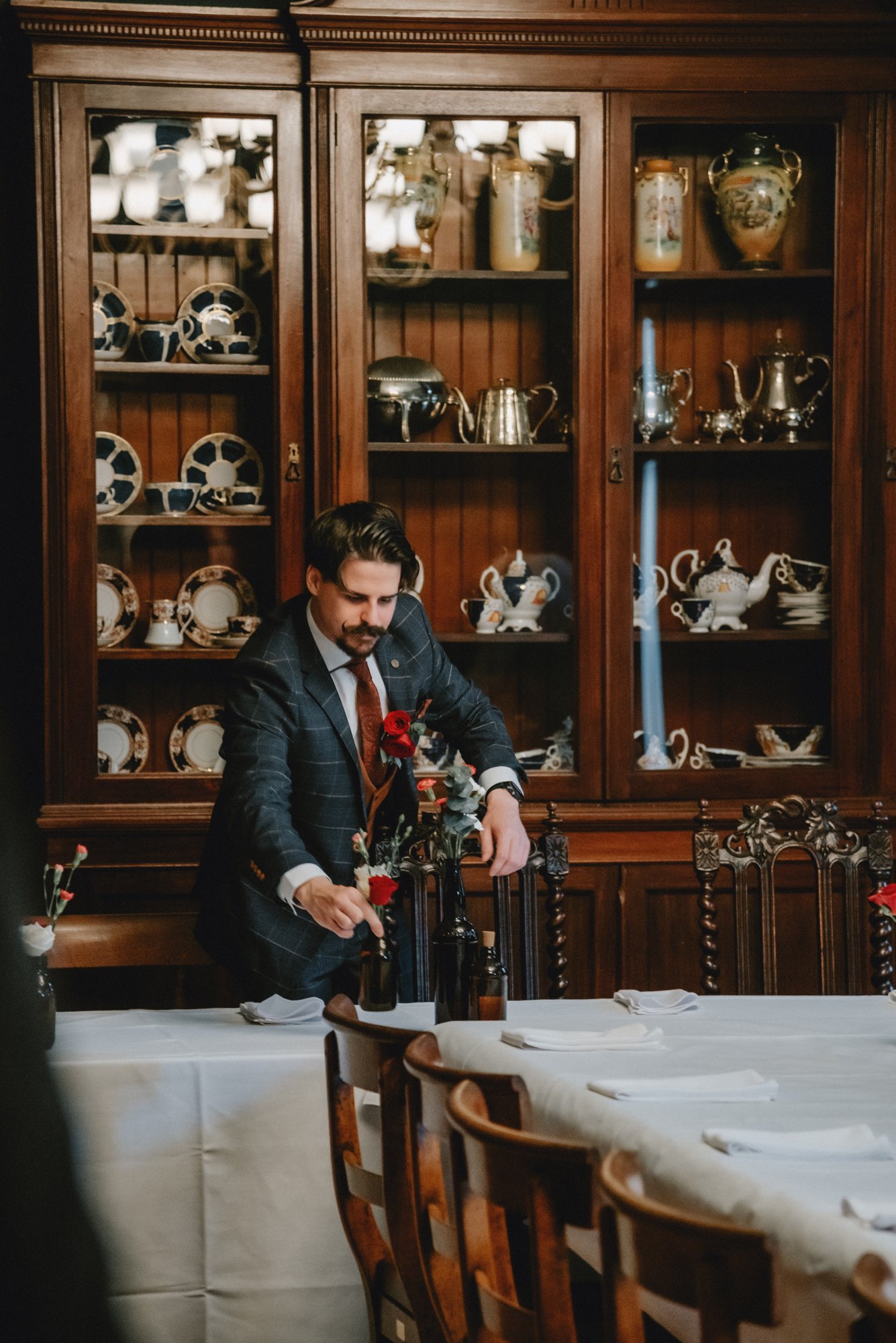 A man in a dark plaid suit arranging flowers on a dining table in a formal dining room with a glass-fronted wooden china cabinet behind him.