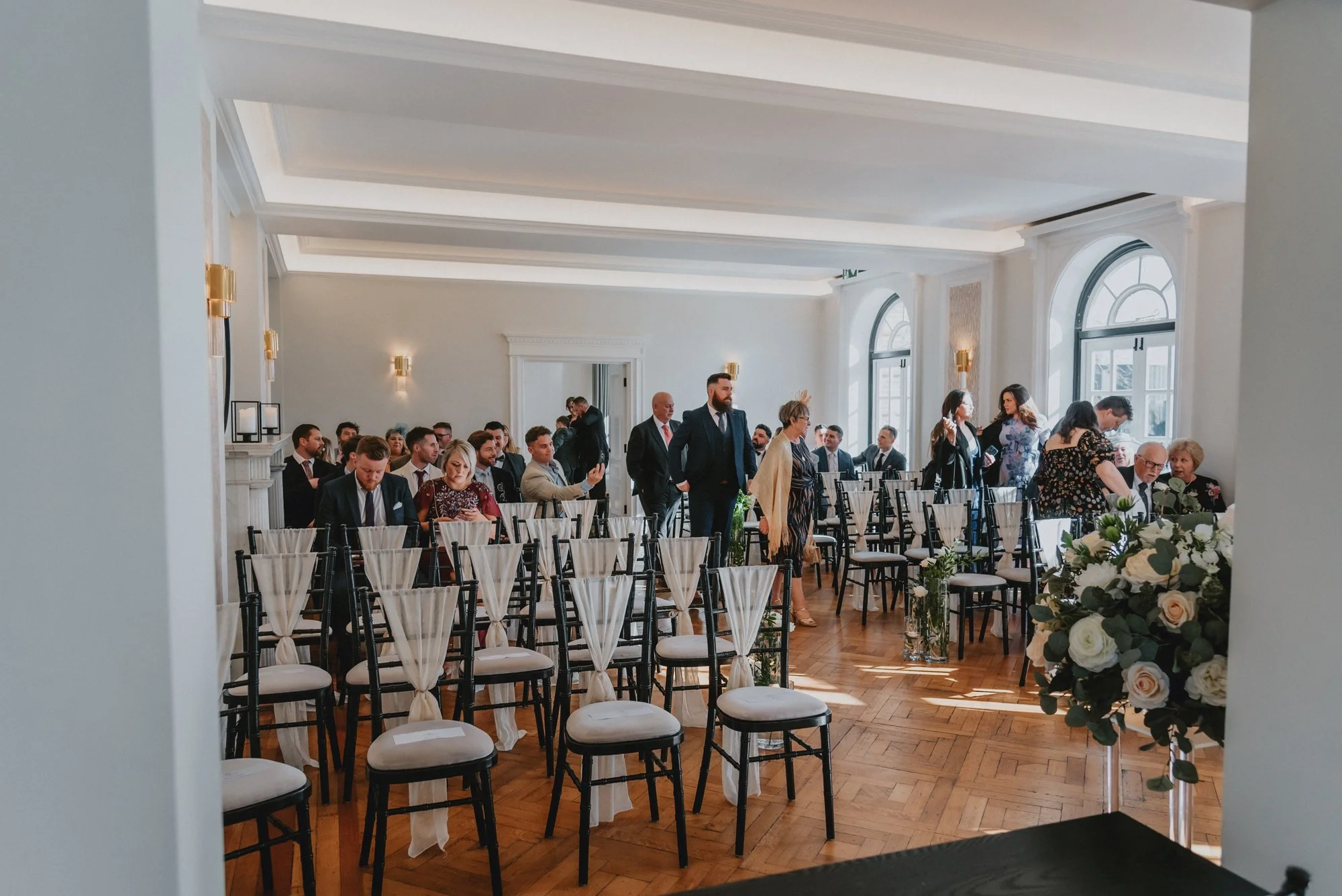 Guests seated in a bright, elegant banquet hall with white walls and large arched windows, some standing and conversing, dressed in formal attire for a wedding or similar event.