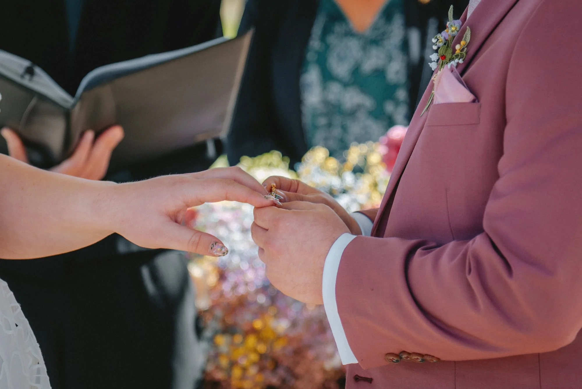 A person in a pink suit exchanging wedding rings with another person at an outdoor wedding ceremony.