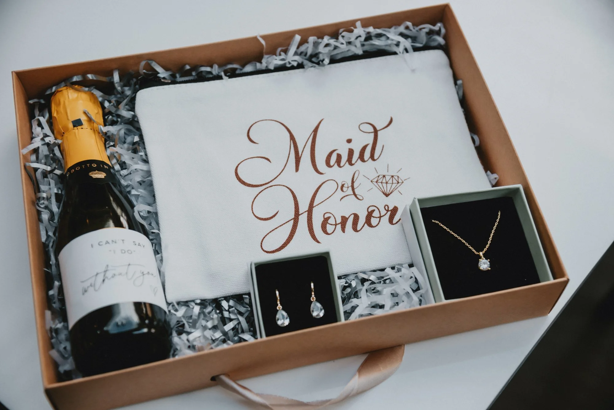 Gift box containing a bottle of champagne, a white cloth with 'Maid of Honor' written on it, a pair of teardrop earrings, and a necklace with a diamond pendant.