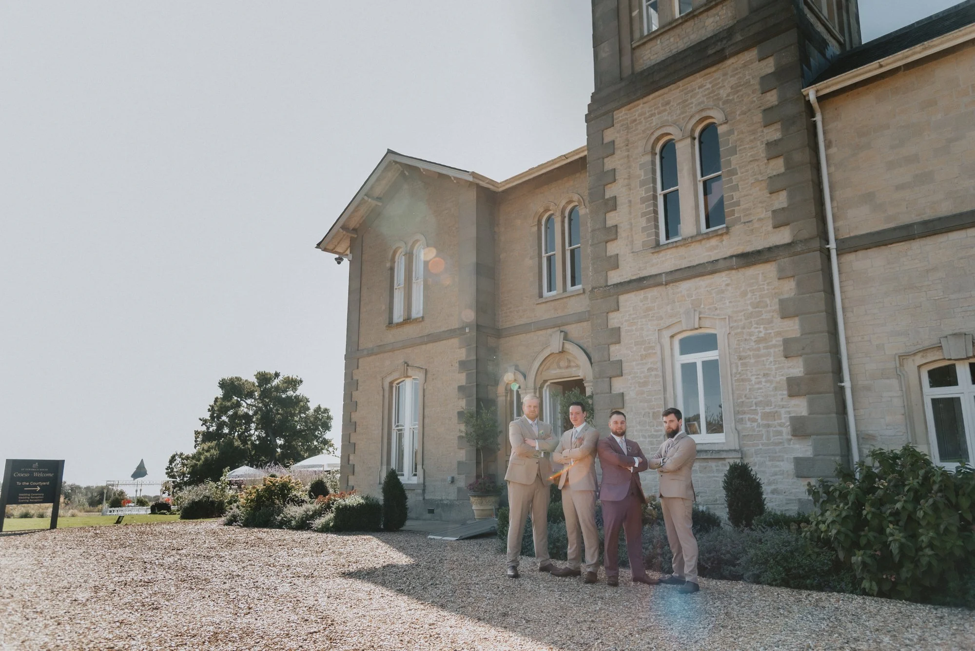 Four men dressed in suits standing outside a large stone building with arched windows, near a garden with bushes and a gravel path.