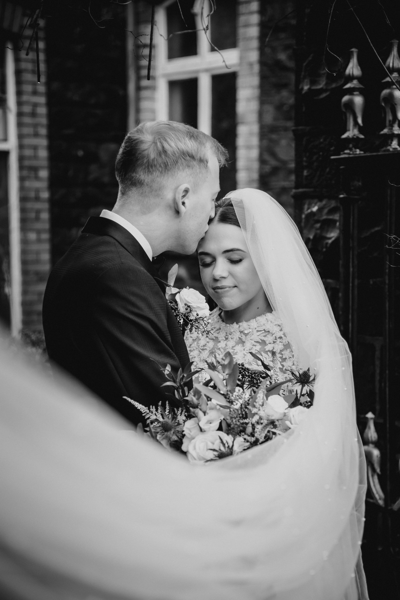 A black and white photo of a bride and groom in an embrace, with the groom pressing his forehead against the bride's forehead. The bride is smiling softly, holding a bouquet of flowers. They are outside, near a brick building with a window in the bac