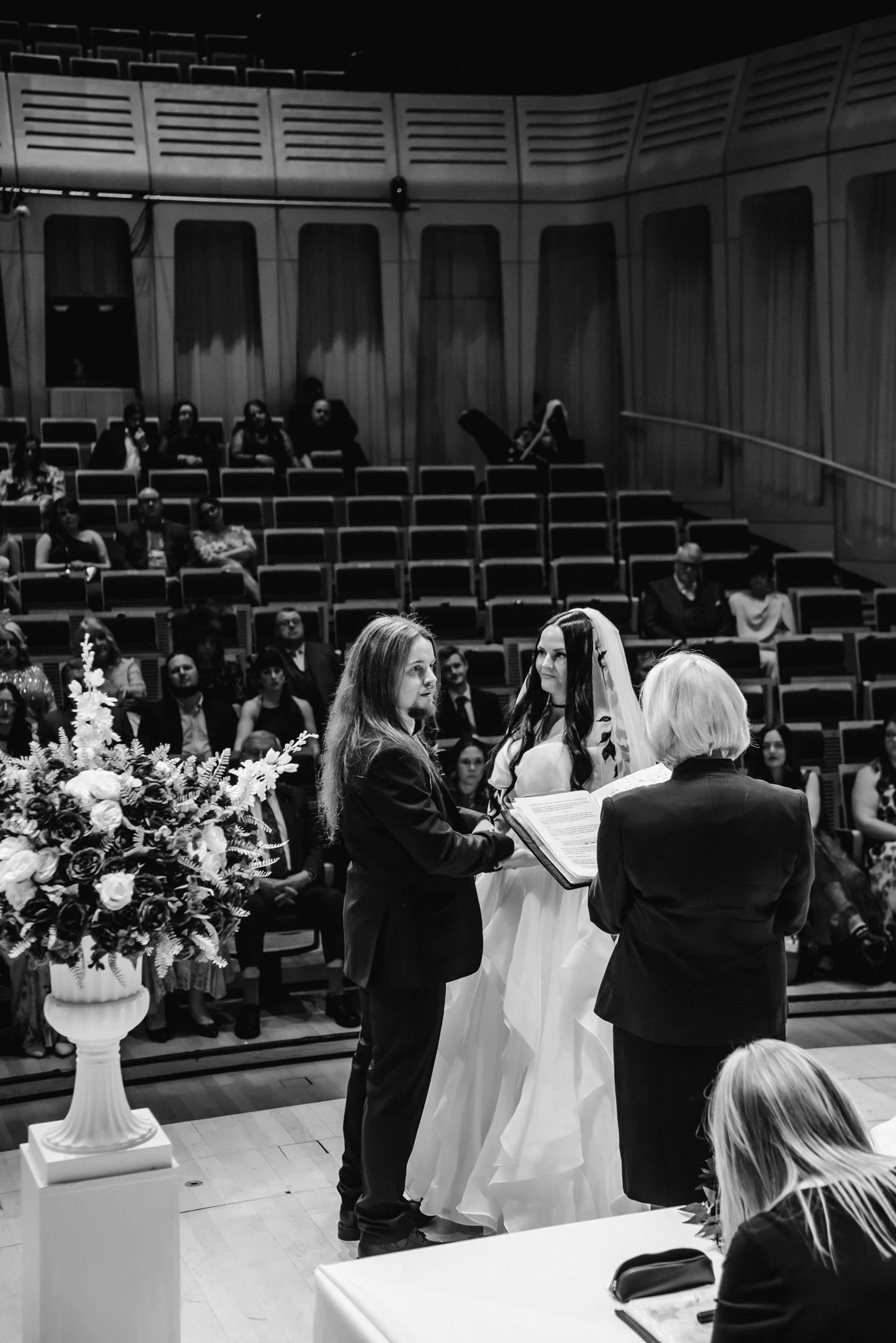 A wedding ceremony with a bride in a white dress and veil, a groom in a suit, and an officiant holding a book, indoors with seated guests in the background.