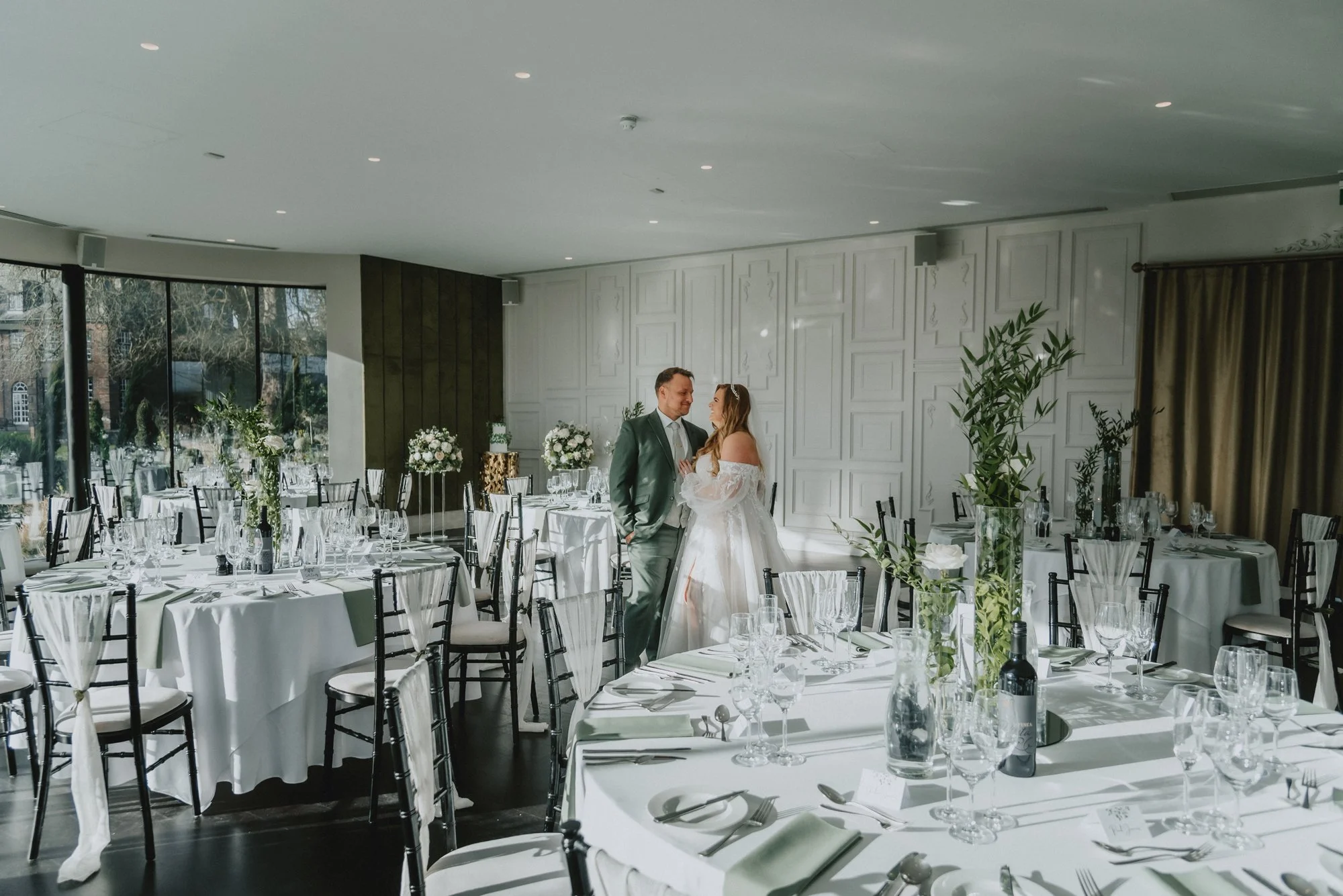 A bride and groom stand close to each other in a decorated wedding reception hall, surrounded by round tables with white tablecloths, glassware, and floral centerpieces.