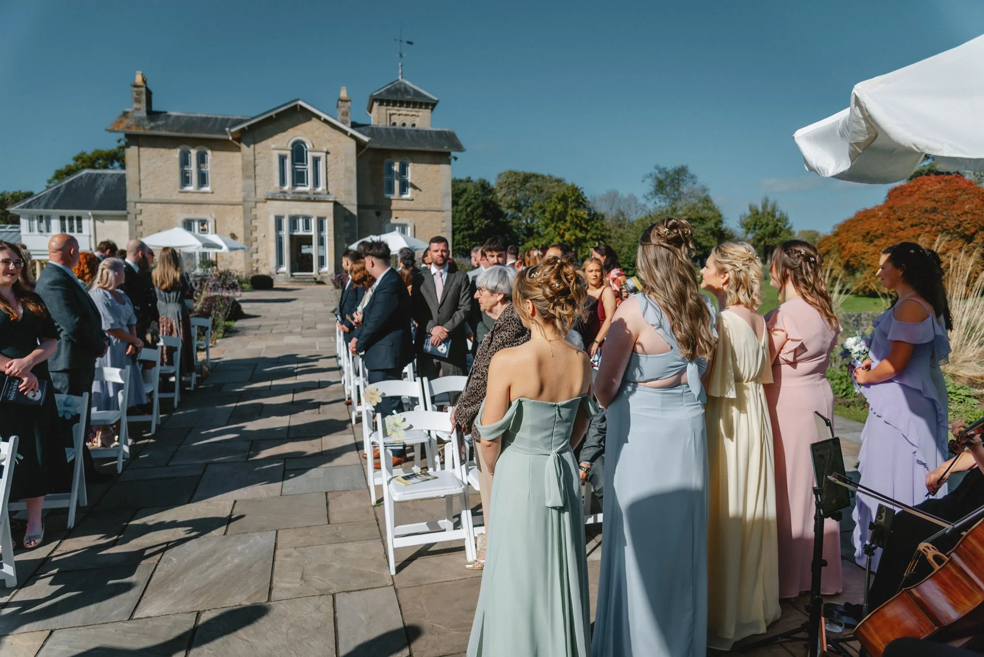 Outdoor wedding ceremony with guests in formal attire, facing a large stone house on a sunny day.