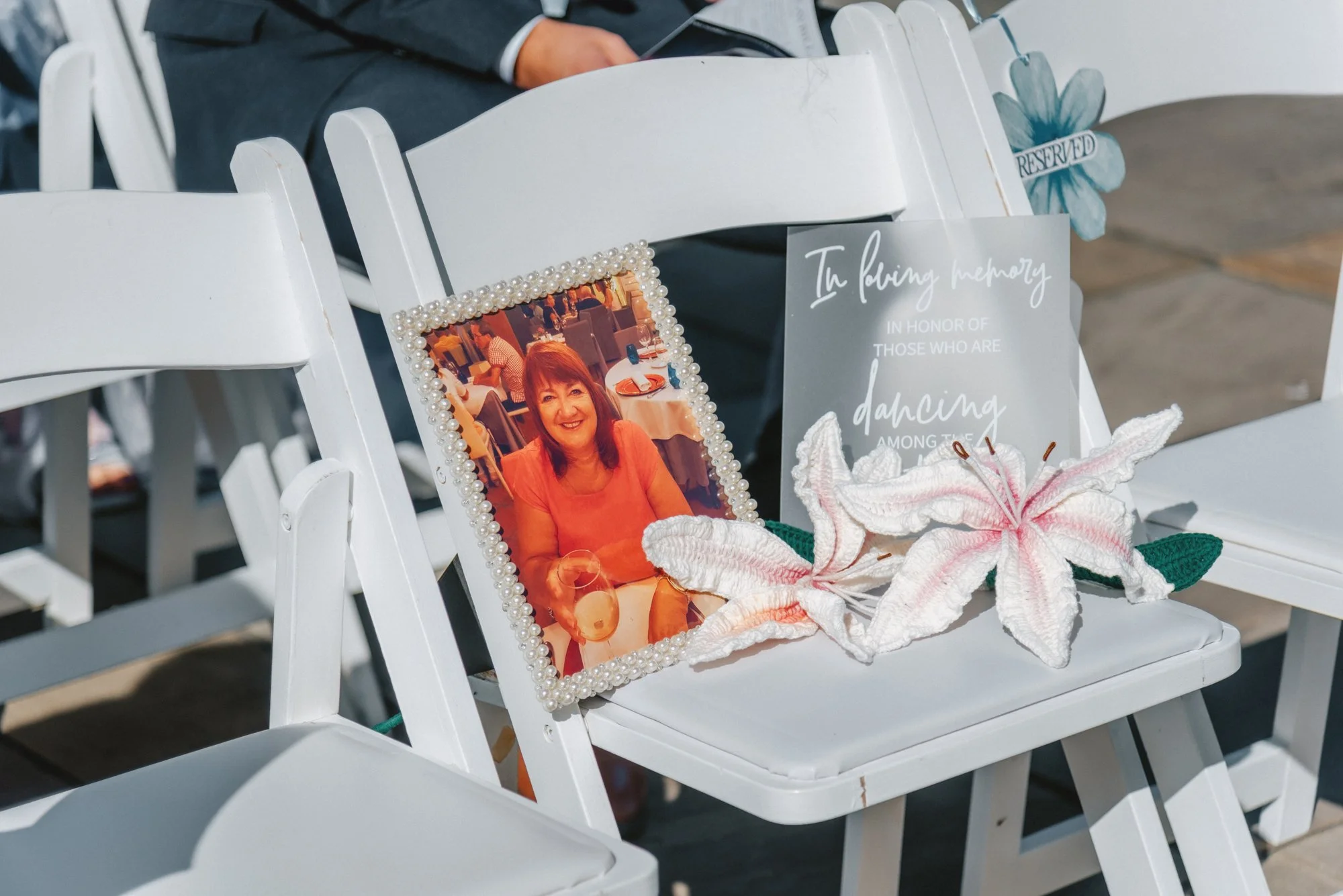 A framed photo of a woman smiling, sitting at a table in a restaurant, decorated with a pearl border, placed on a white chair. Next to the photo is a memorial card with white flowers, lilies, and a printed message in memory of those who are dancing a