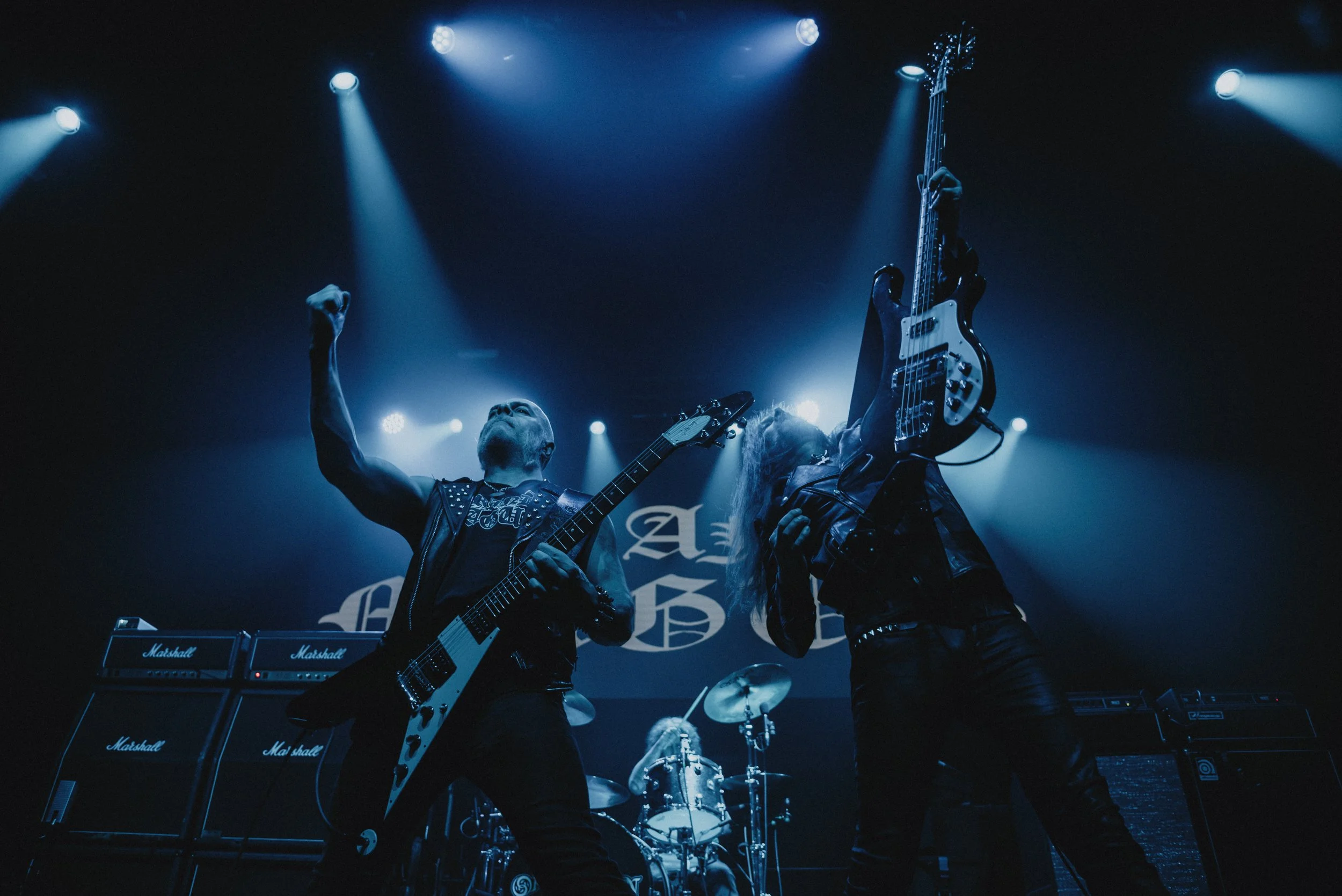 Two guitarists and a drummer performing on stage during a rock concert with blue lighting and a backdrop.