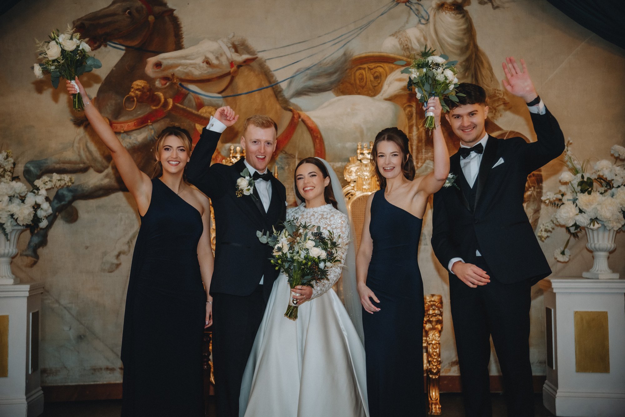Group of five people at a wedding, with the bride holding a bouquet in the center, three women in navy blue dresses, and two men in black tuxedos, all smiling and raising flowers or hands in celebration, standing in front of a mural of horses.