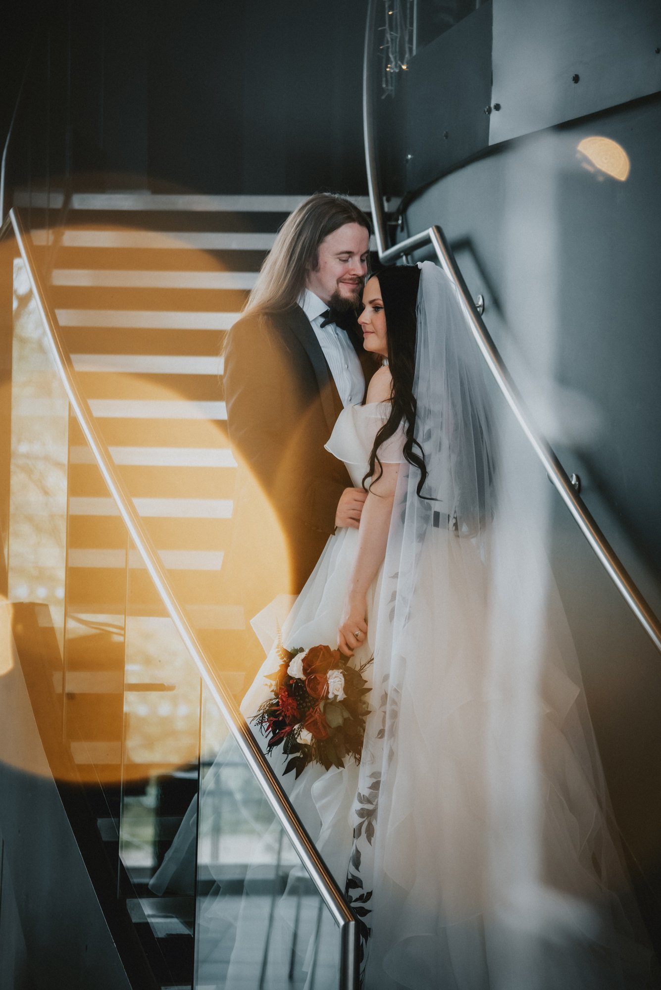 Bride and groom sharing an intimate moment on a staircase, with the bride holding a bouquet of flowers and the groom in a tuxedo, reflected in a glass surface.