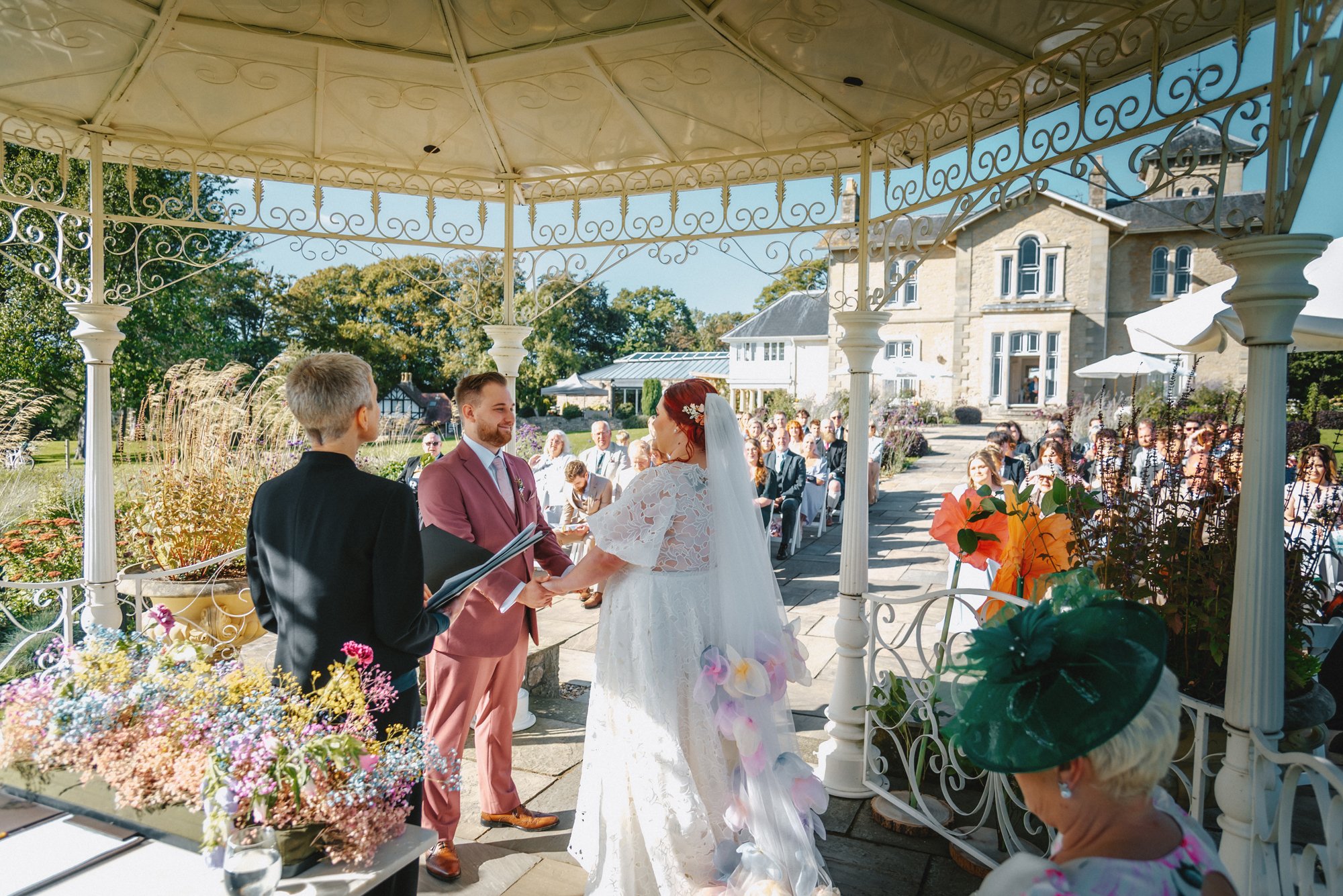 A wedding ceremony taking place outdoors under a decorative gazebo with the bride and groom holding hands and exchanging vows, surrounded by seated guests and a large house in the background.