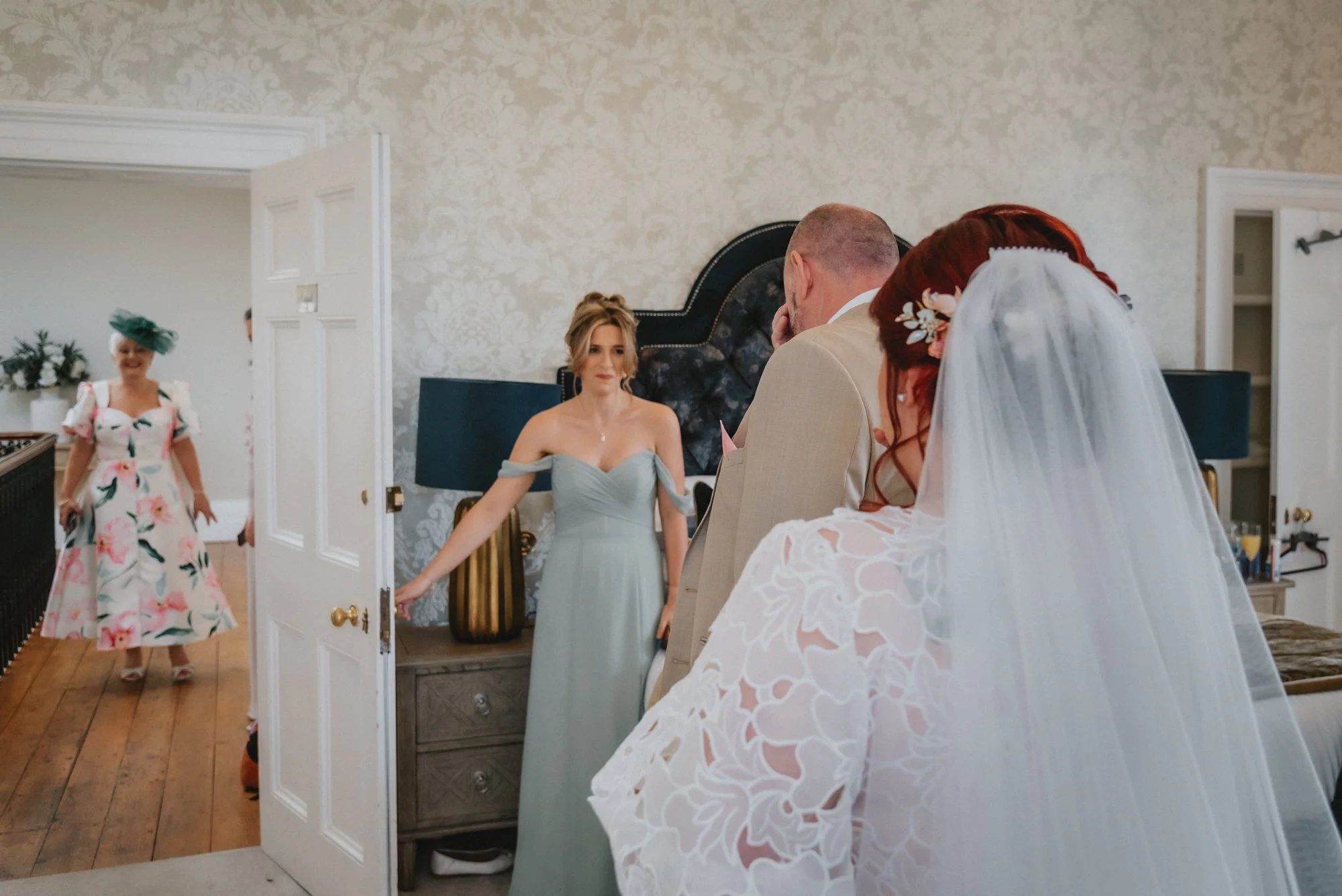 A bride with a white veil and lace dress is standing with a person and a man, as another woman in a floral dress and hat enters through an open door in a bedroom
