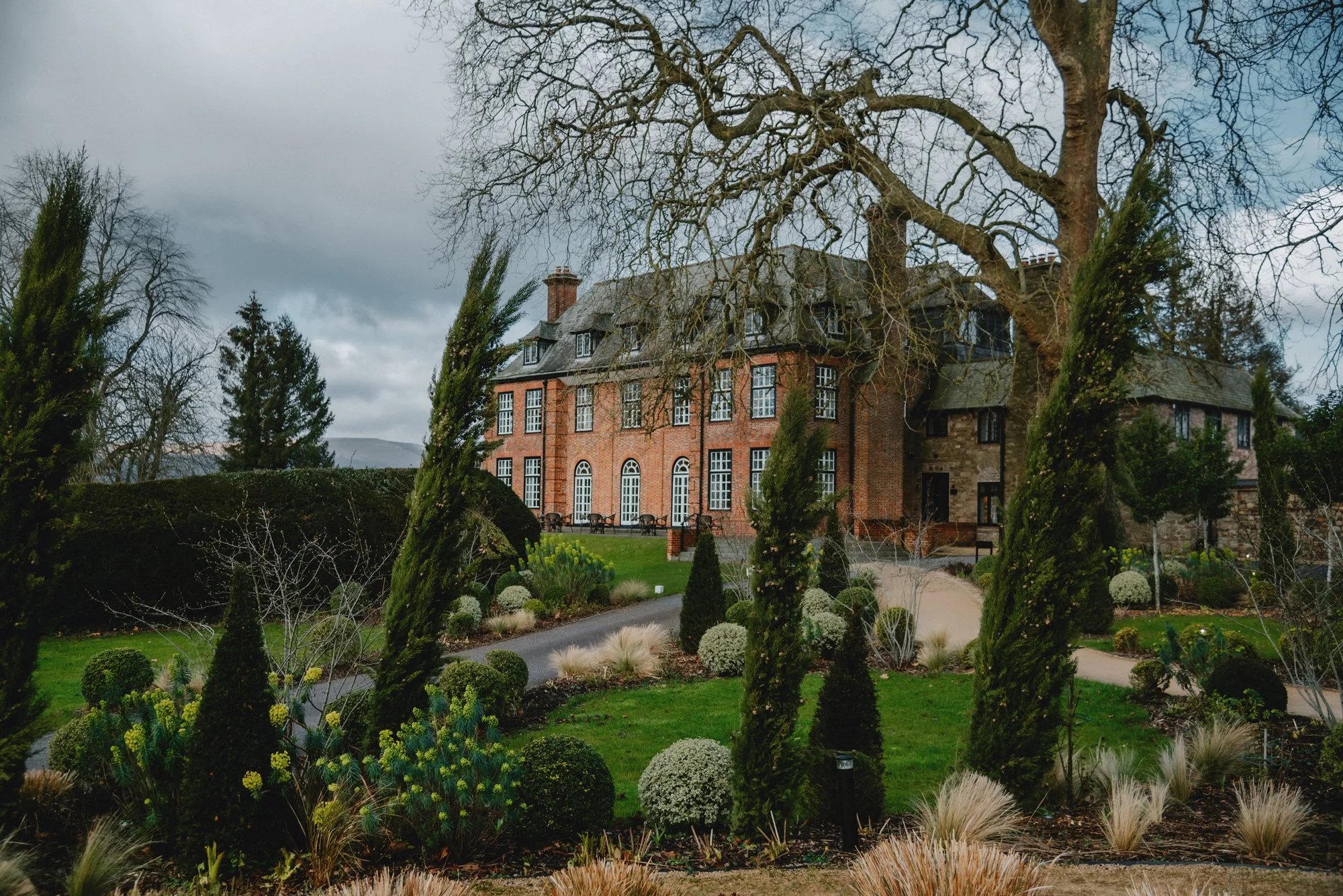 A large brick mansion surrounded by landscaped garden with tall cypress trees, bushes, and a winding pathway