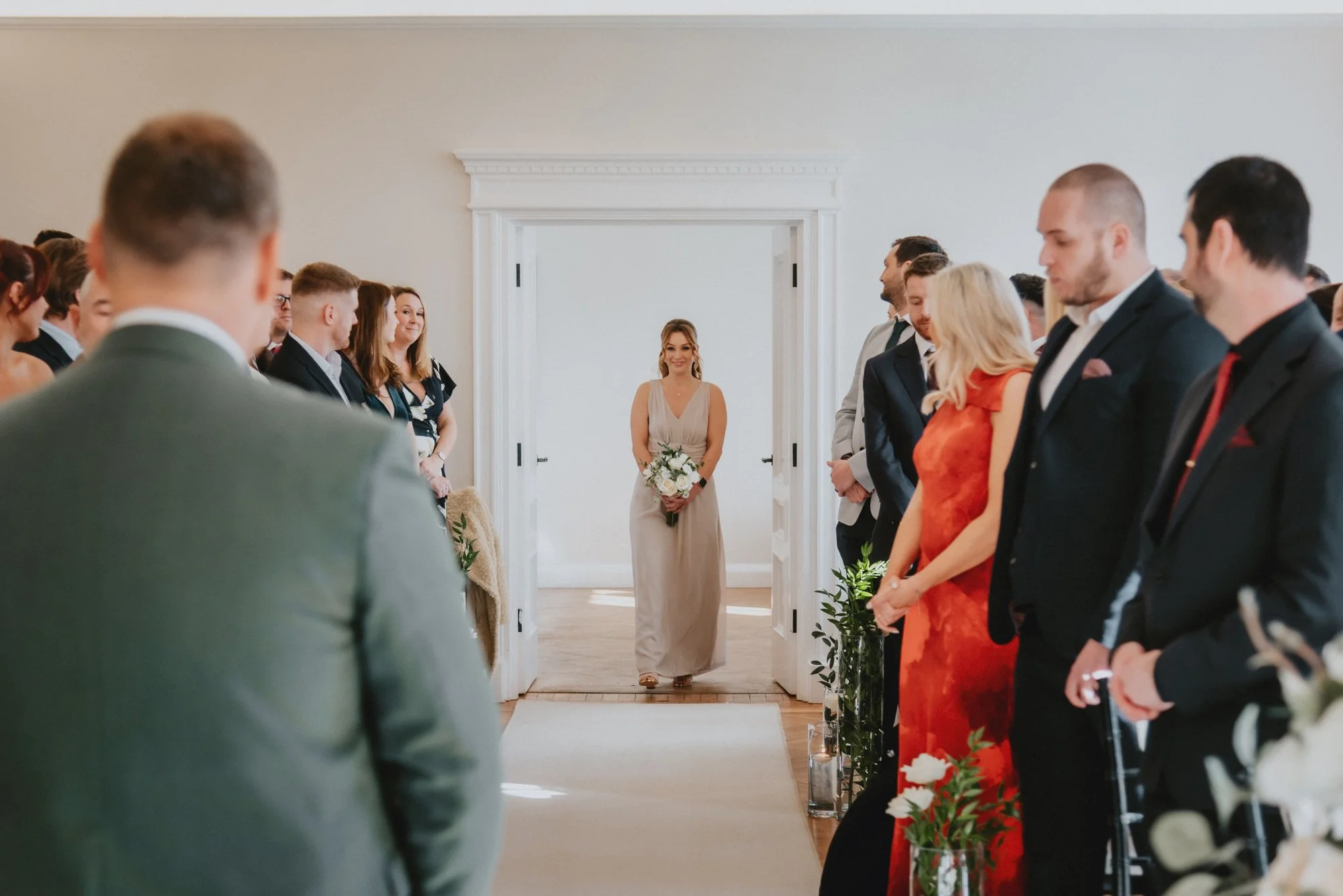 A wedding ceremony with a bride walking down the aisle toward the groom, who is waiting on the other end, surrounded by seated guests in formal attire.
