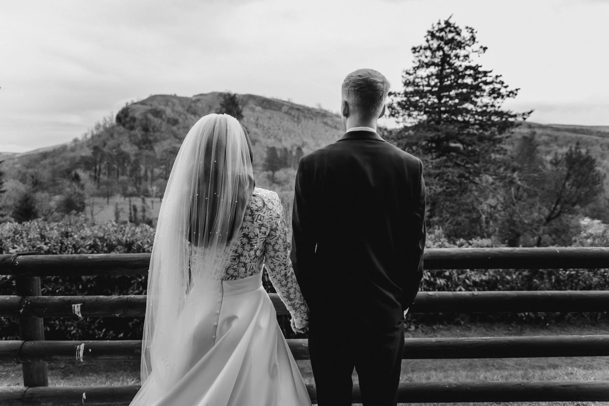 A black-and-white photo of a bride and groom standing side by side, facing away, looking at a scenic landscape with hills and trees.