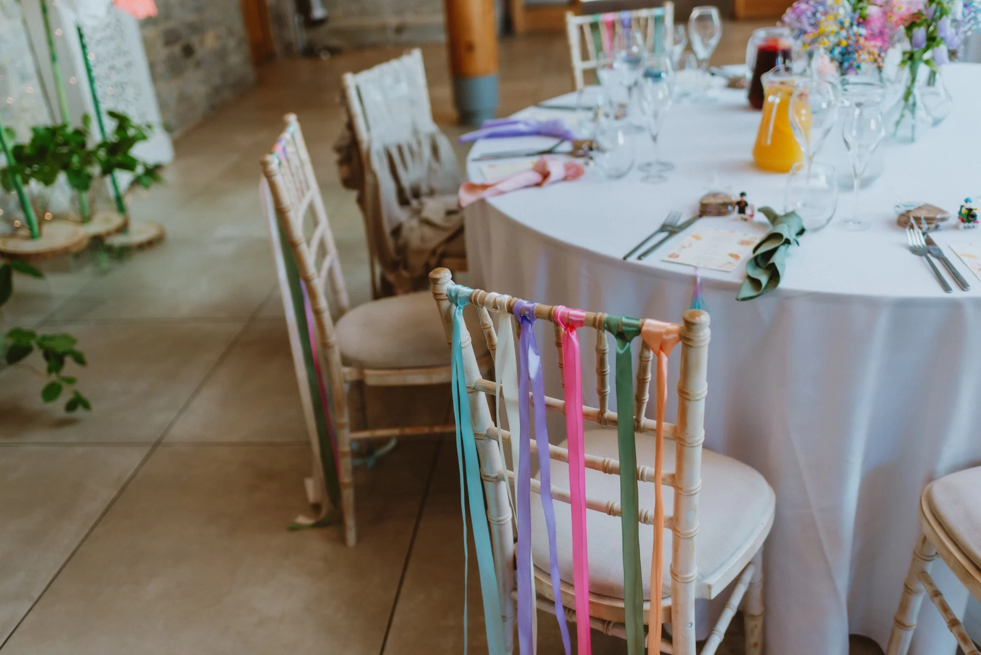 Decorated chairs with colorful ribbons at a wedding or party table setting with flowers, glassware, and dishes on a white tablecloth in a rustic venue.
