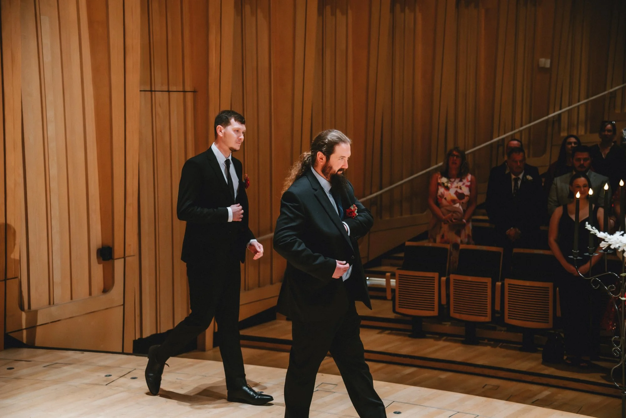 Two men in formal suits walking in an event hall with wood-paneled walls, surrounded by seated guests.