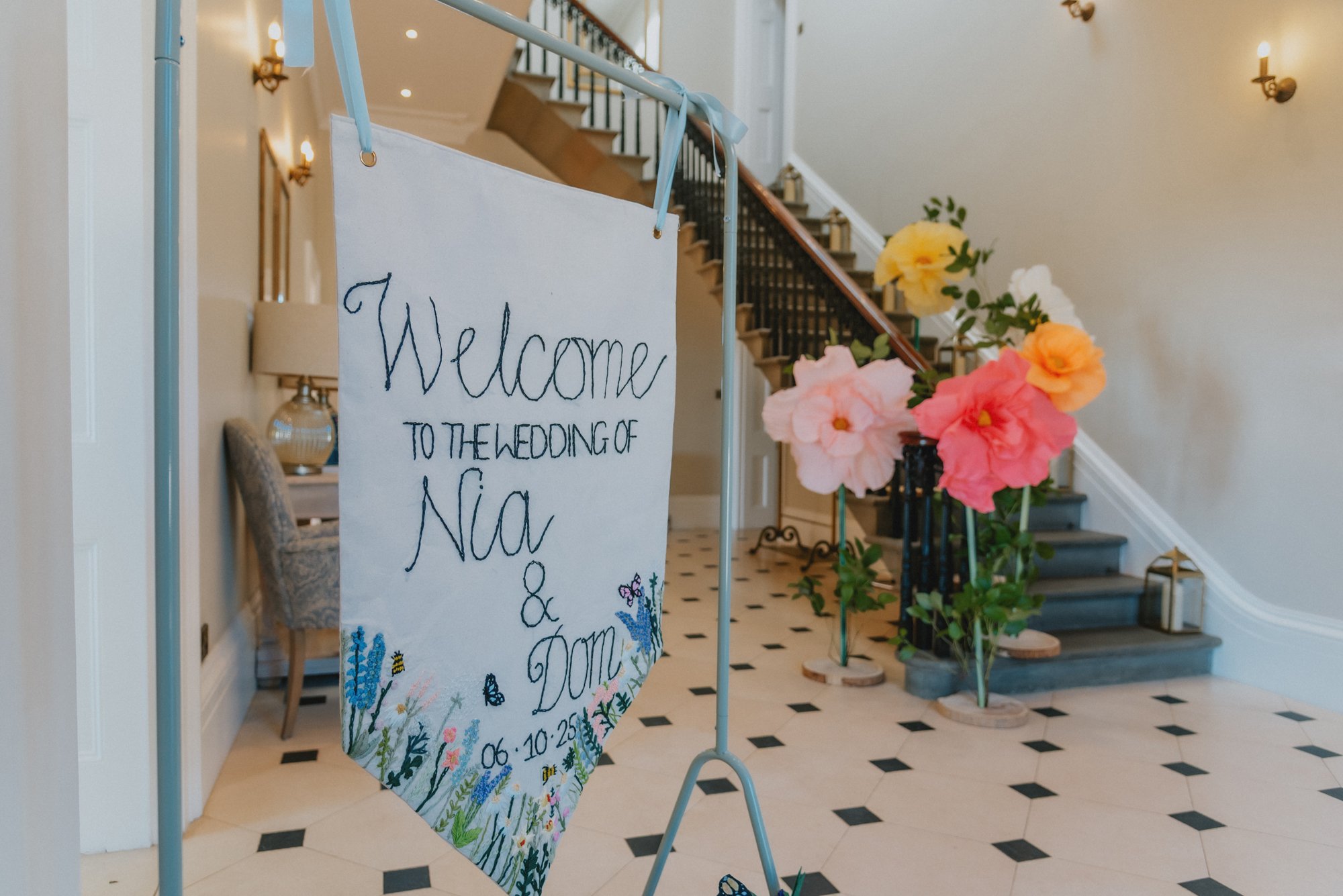 Welcome banner for the wedding of Nia and Dom, displayed near pink, yellow, and peach flowers at the entrance of an indoor venue with stairs and black-and-white tiled floor.