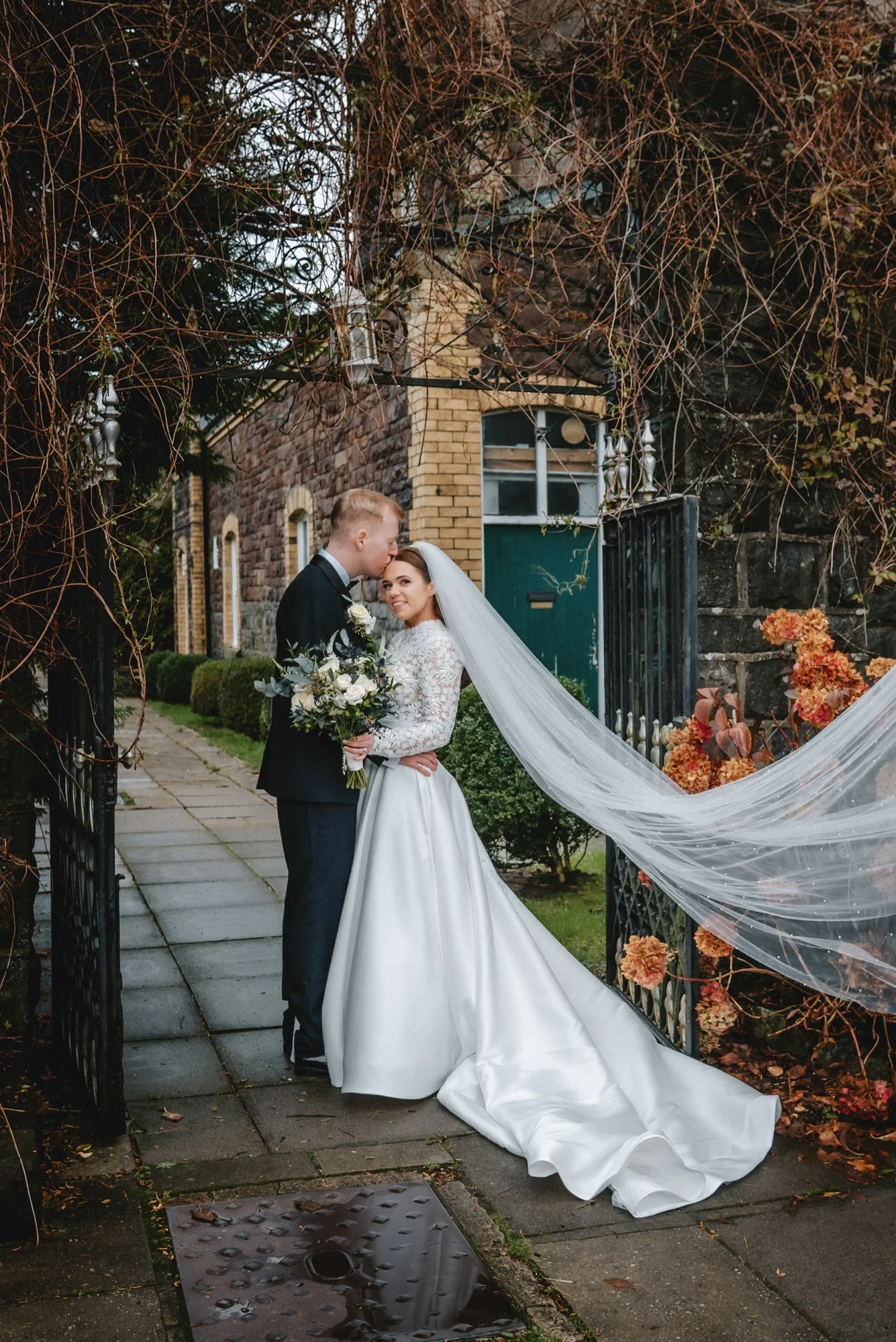 A bride and groom standing close together outdoors on a wedding day, with the groom kissing the bride on her forehead. The bride is smiling, holding a bouquet of flowers, wearing a long-sleeved lace wedding dress and veil. The groom is dressed in a b