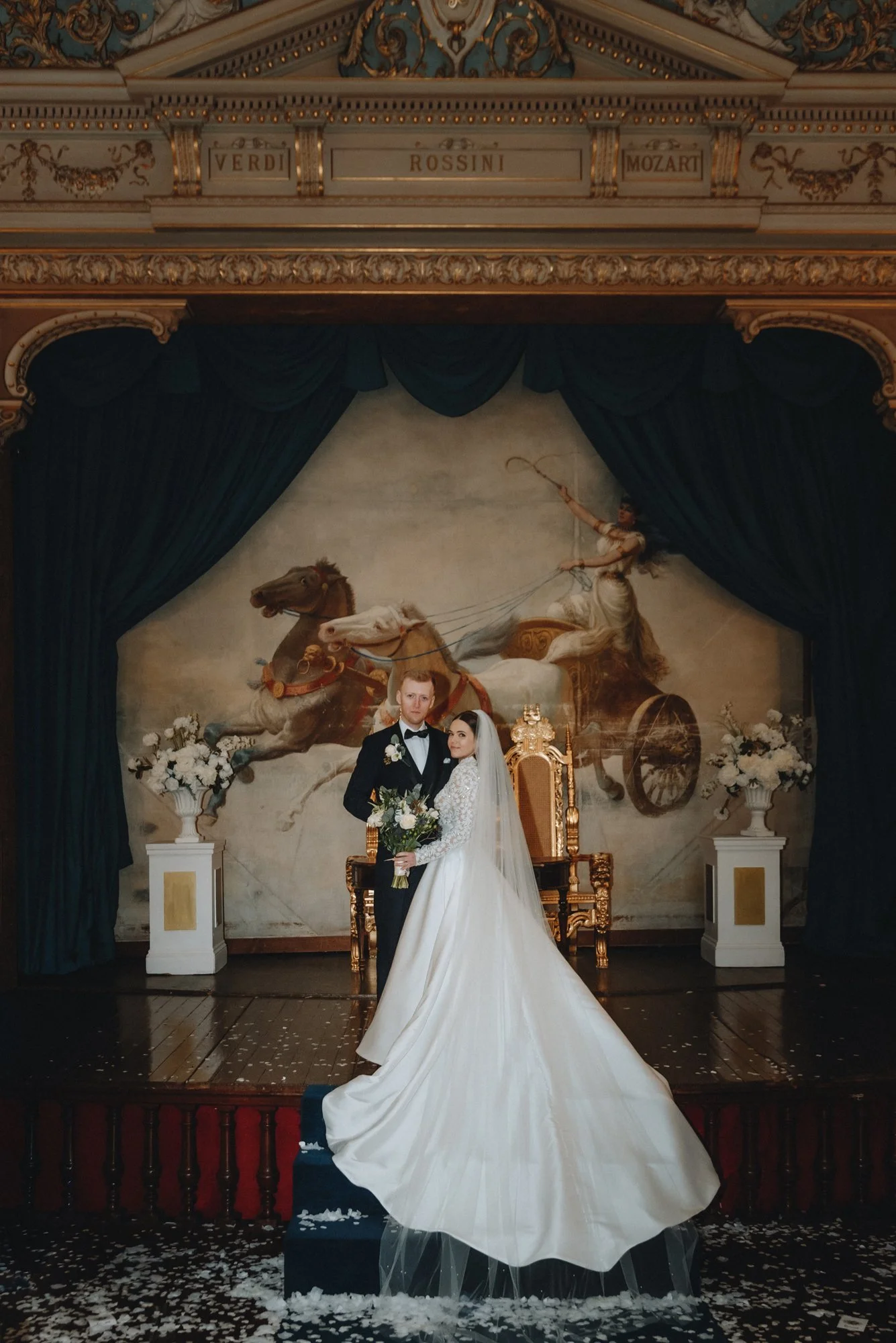 A bride and groom standing on a small stage, posing for a wedding photo in front of a royal throne. The bride wears a white wedding dress with a long train and veil, holding a bouquet of flowers. The groom is dressed in a black tuxedo. Behind them, t