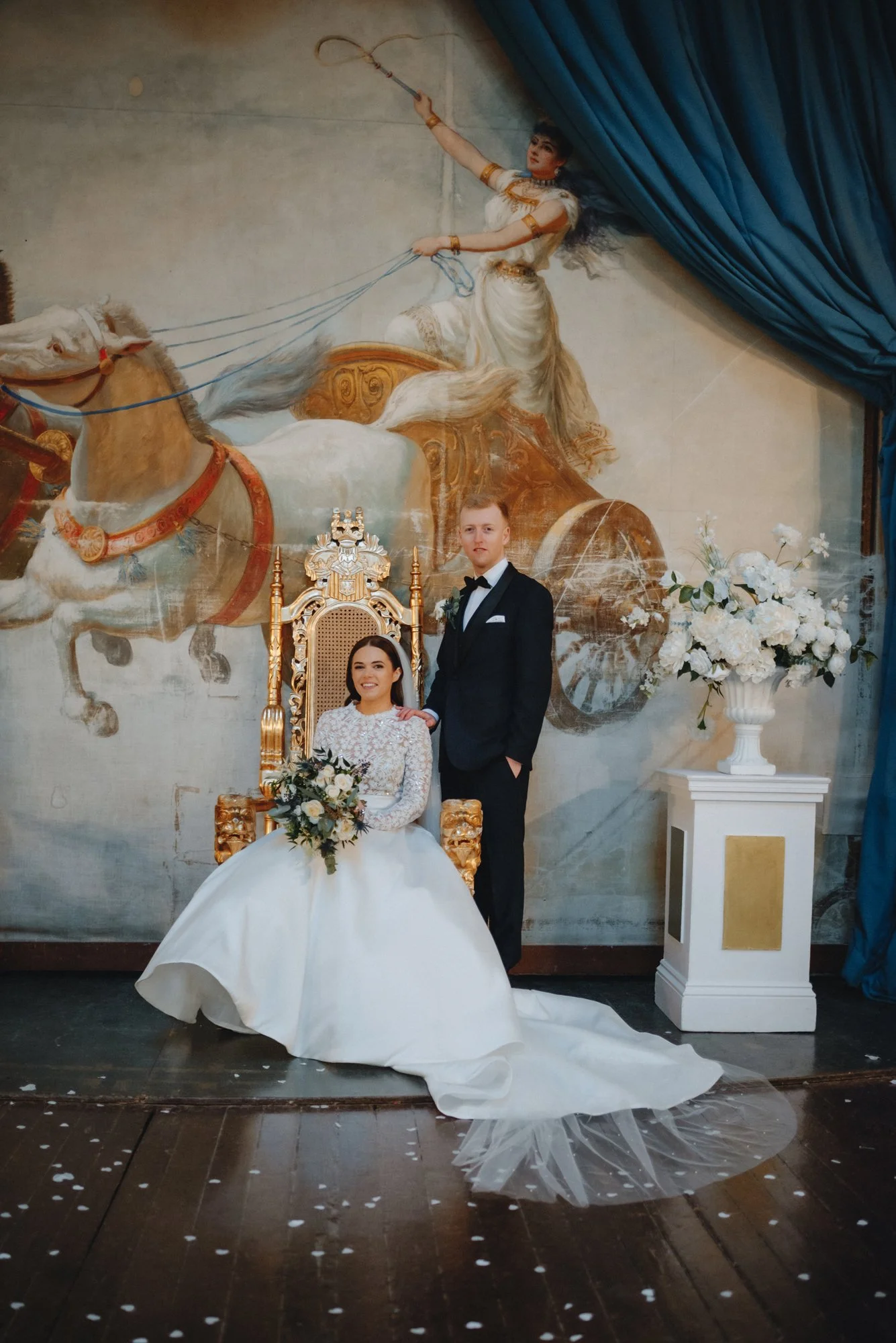 A bride in a white wedding dress sitting on a golden throne-like chair, holding a bouquet, and a groom in a black tuxedo standing beside her with his hand on her shoulder. Behind them is a large mural of a woman in a chariot drawn by a horse.