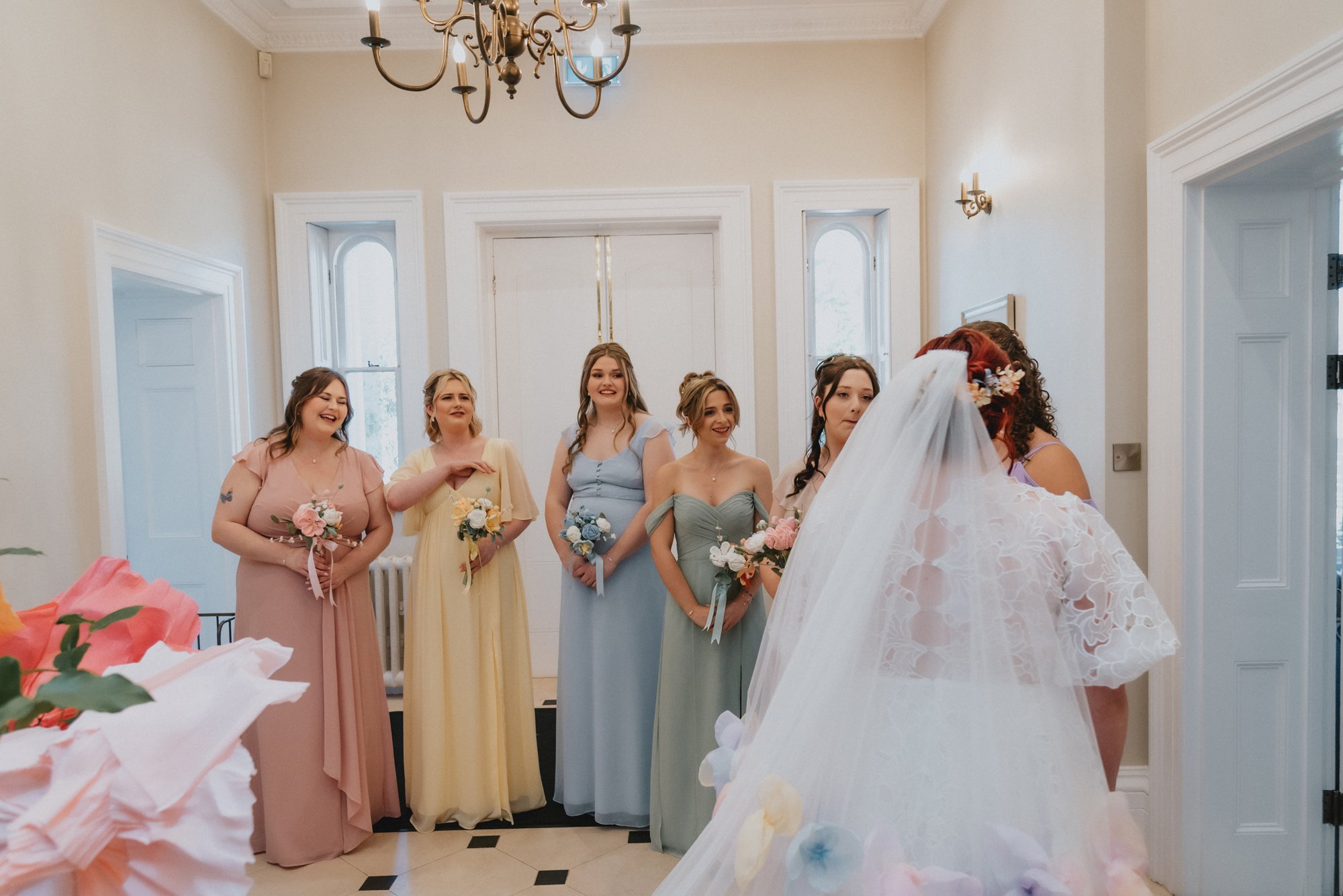 A bride, with red hair and a veil, facing five bridesmaids holding bouquets, in a bright room with white walls, high ceilings, and a chandelier.