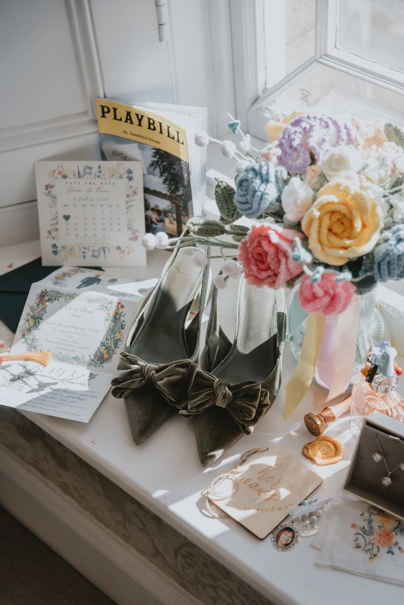 A collection of decorative and functional items on a table, including a bouquet of colorful knitted flowers, a pair of gray shoes with bows, greeting cards, a small jewelry box with jewelry, a picture frame, and a playbill, all near a window with nat