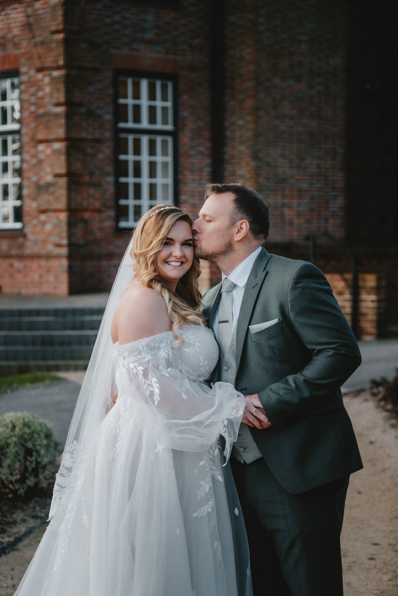 A newlywed couple standing outdoors in front of a brick building, with the groom kissing the bride's forehead as she smiles.