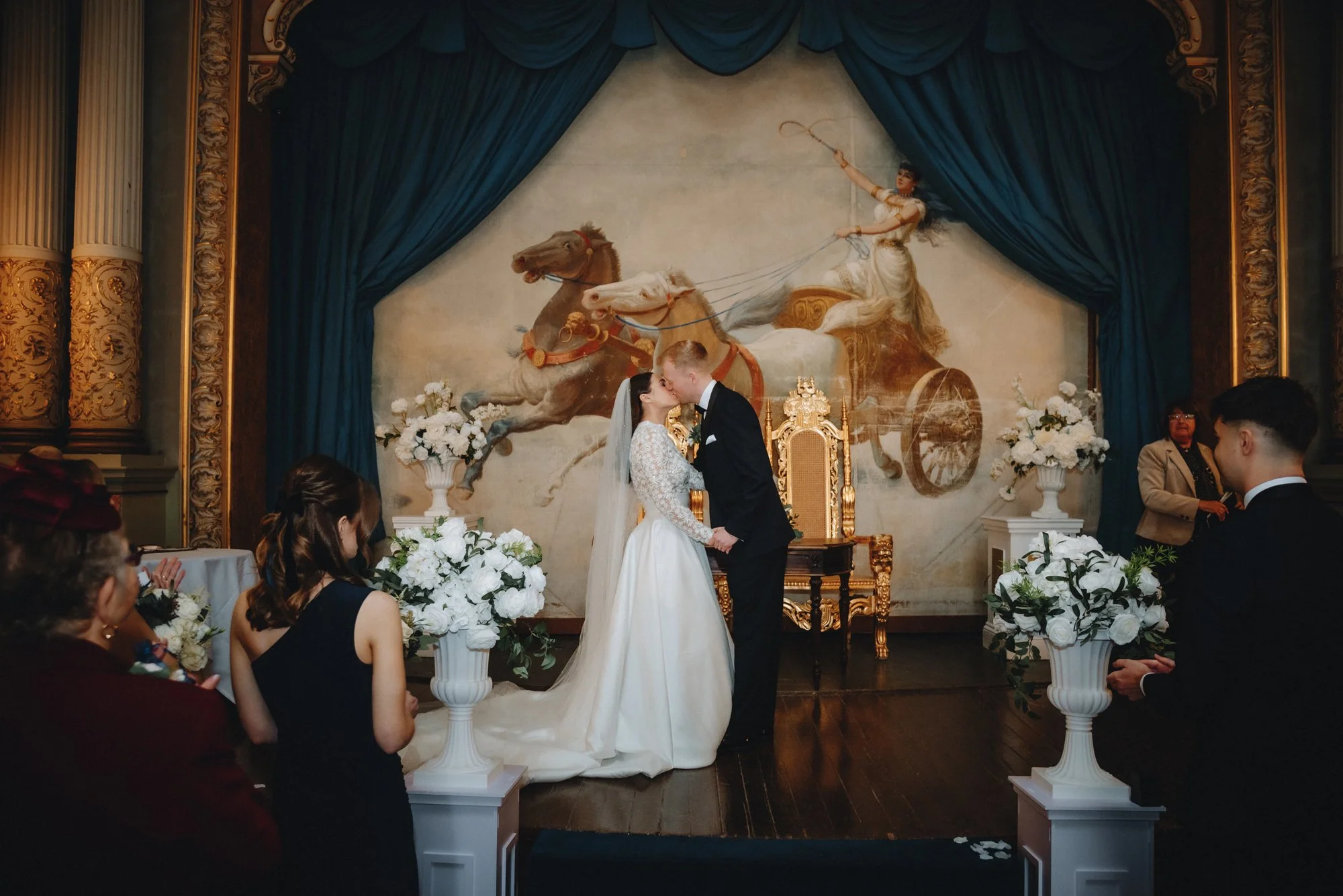 A wedding ceremony taking place indoors with a couple kissing at the altar. The bride wears a white wedding dress and veil, and the groom in a black tuxedo. The background features a large mural of a classical chariot scene with a woman riding a char