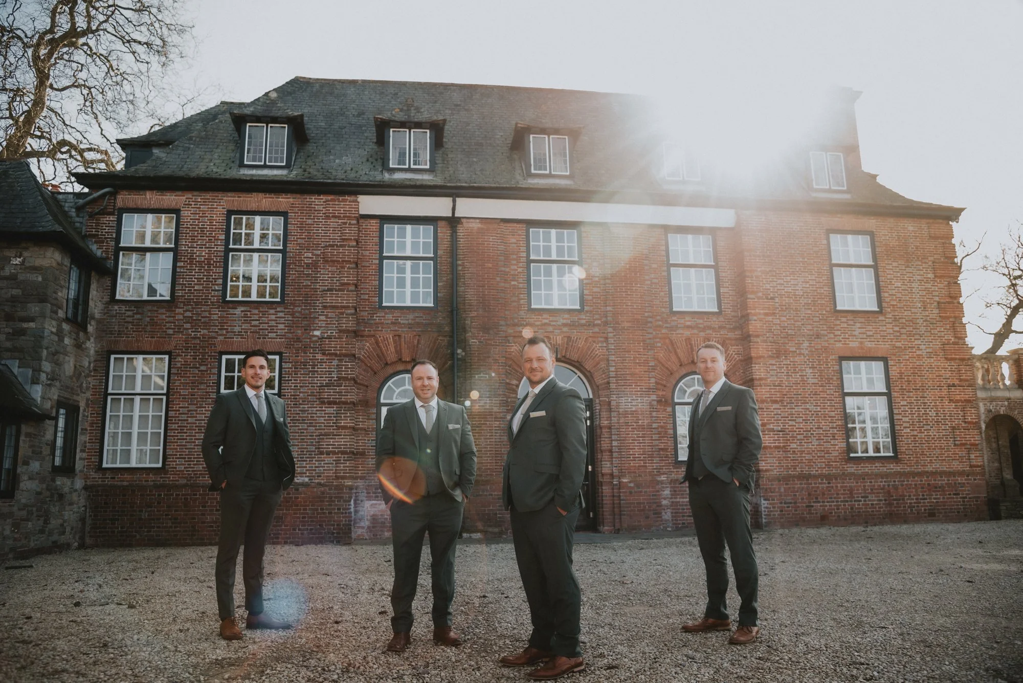 Four men in suits standing outside a large brick building with multiple windows, under sunlight.