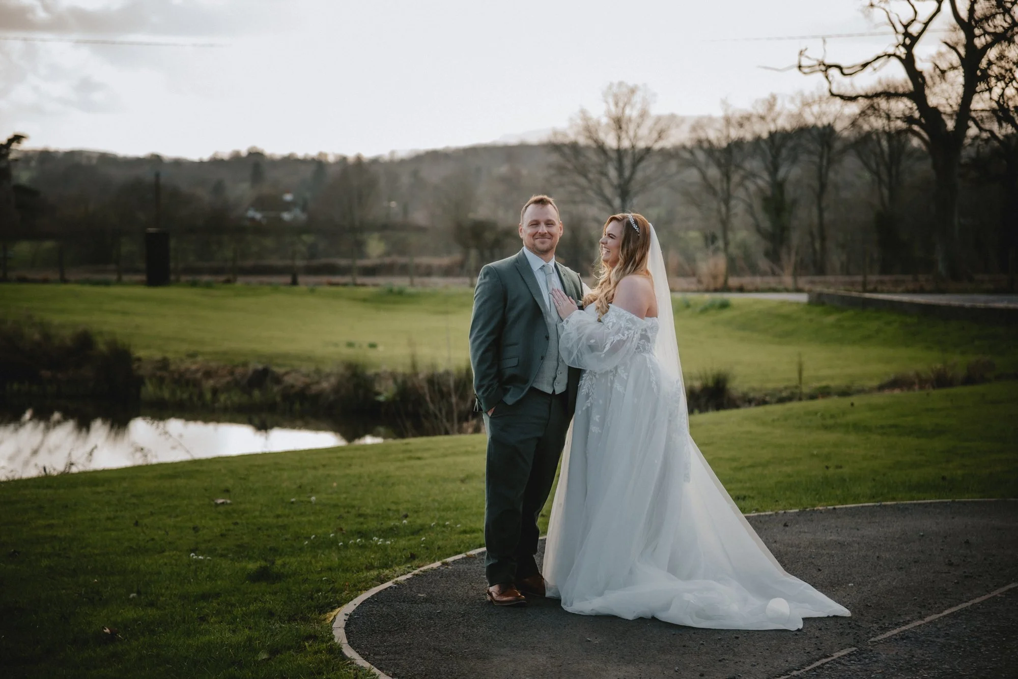 A bride and groom standing outdoors on a paved area during sunset, with a grassy landscape and trees in the background. The bride is wearing a white off-shoulder wedding gown and veil, and the groom is dressed in a suit with a tie.