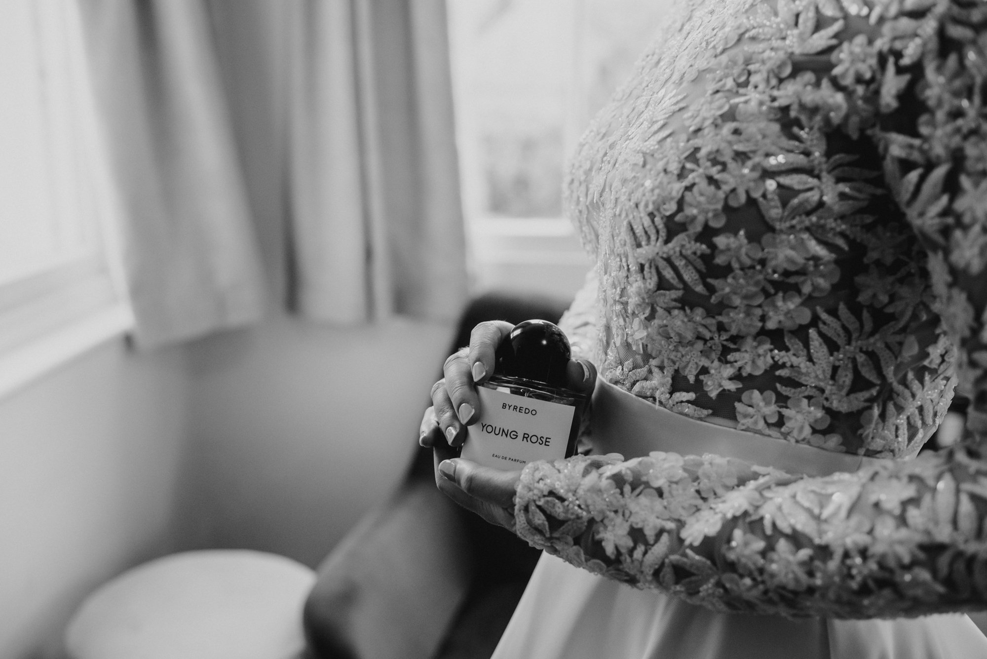 A woman in an embroidered dress holds a perfume bottle labeled "Young Rose" by Byredo in a softly lit room with a window and curtains in the background.