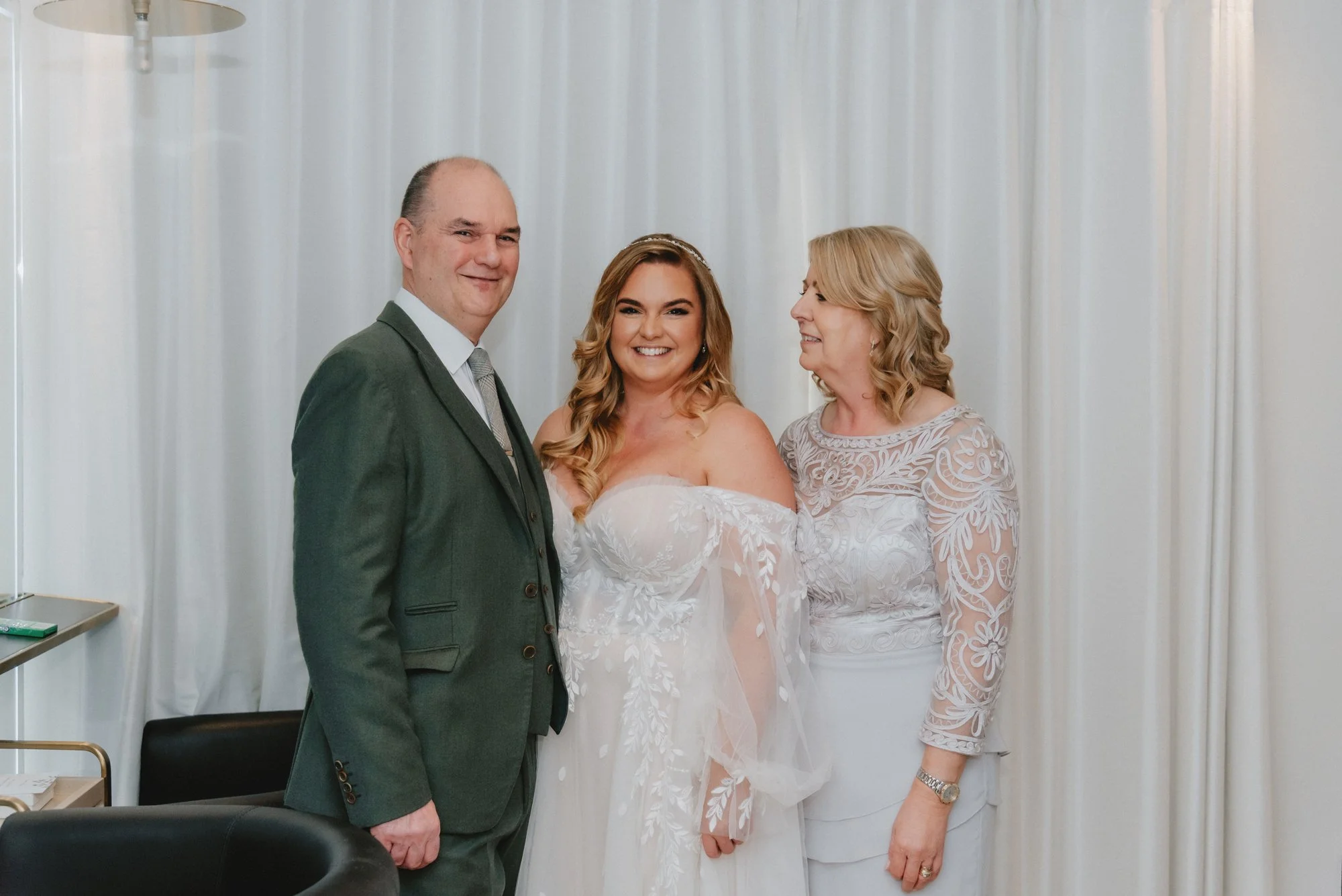 A wedding scene with a bride and two older adults, likely her parents, standing together inside a room with white curtains. The bride is wearing a white off-the-shoulder wedding dress, and the woman on the right is wearing a white lace dress. The man